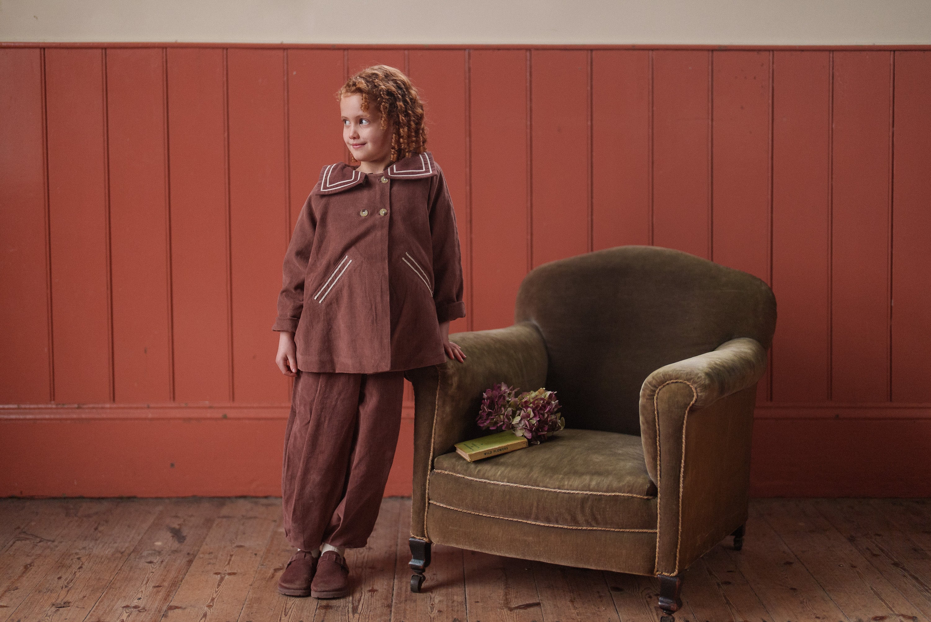 A young child wearing vintage style velvet trousers and matching coat in a warm nutmeg tone, standing next to a green velvet armchair in a cozy wooden room.
