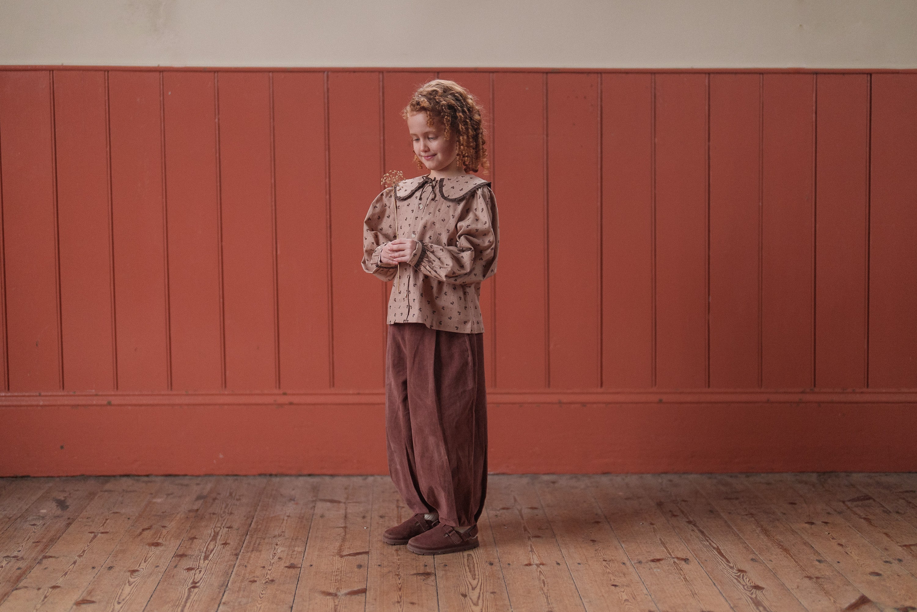 Young girl wearing vintage-inspired girls blouse and loose nutmeg-colored velvet trousers standing on wooden floor against rustic red wall, showcasing sustainable children’s clothing.