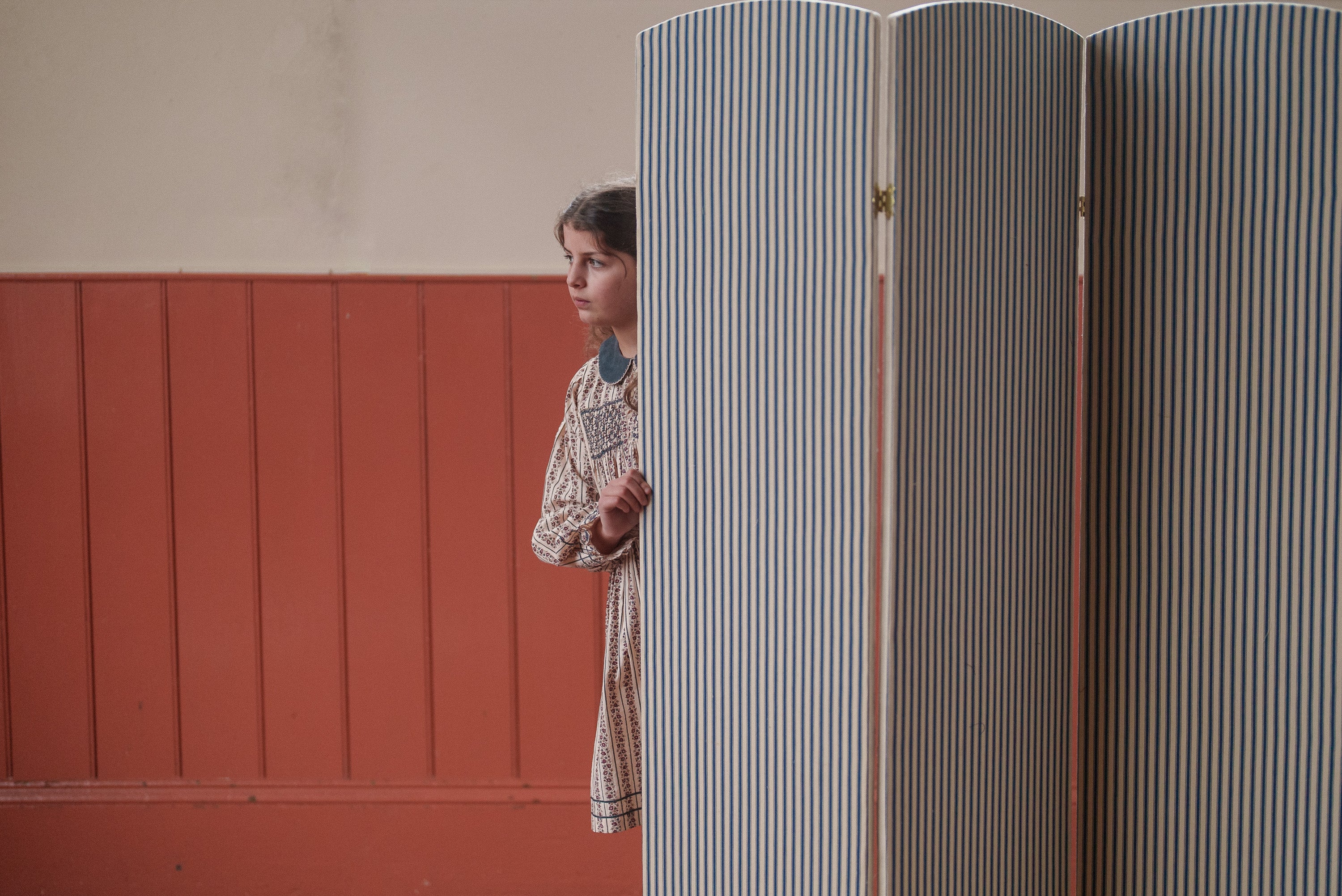 Young girl wearing a vintage style, organic cotton Elizabeth smocked dress with Mallory floral pattern, standing partially behind a blue and white striped room divider in a warm, softly lit space.
