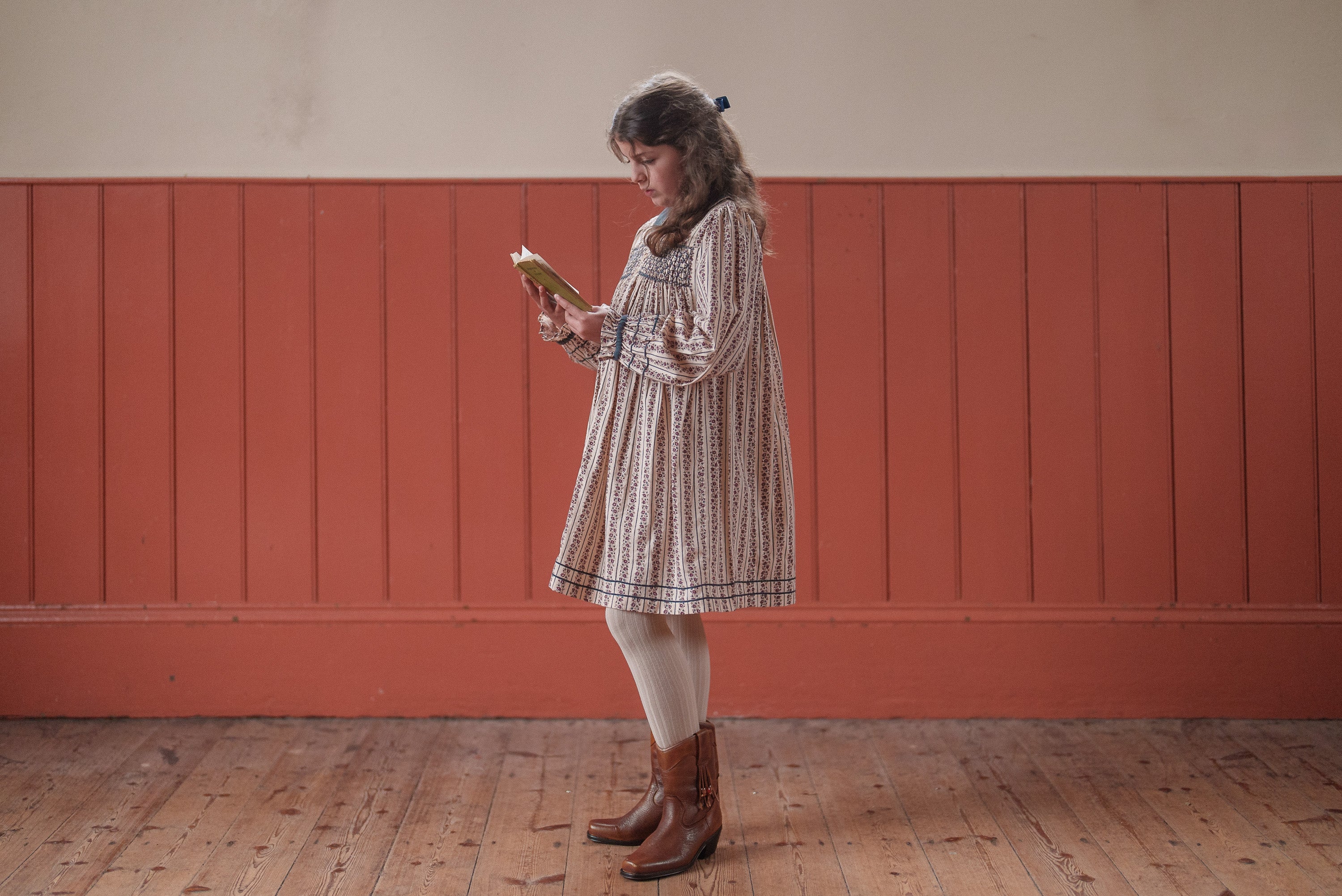 Young girl wearing a vintage style organic cotton dress with smocked details and floral print, standing indoors reading a book, showcasing sustainable children's clothing.