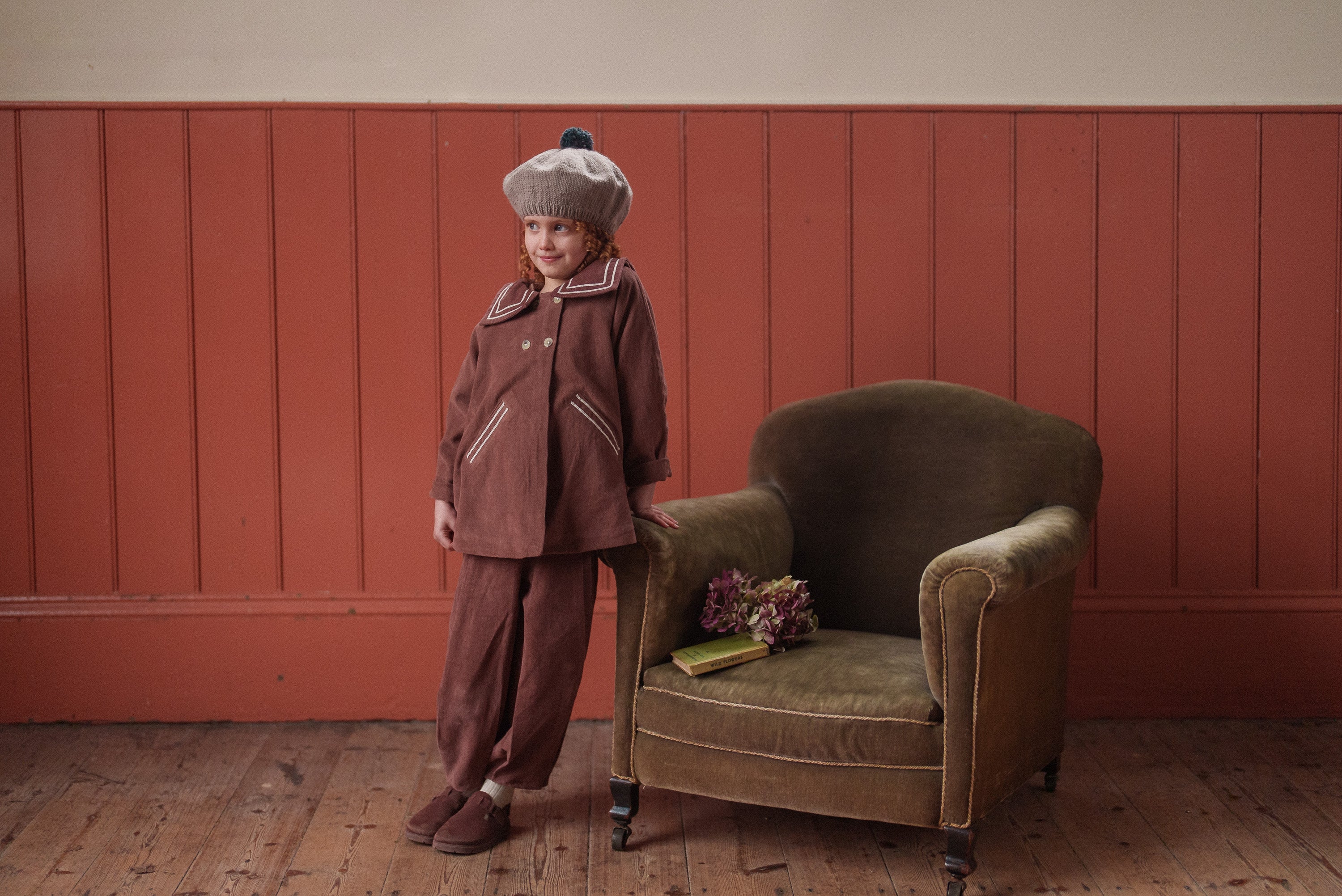 Child wearing vintage style hand-knitted multicolor merino beret and brown traditional children's clothing standing next to an olive green armchair in a warm, natural setting.