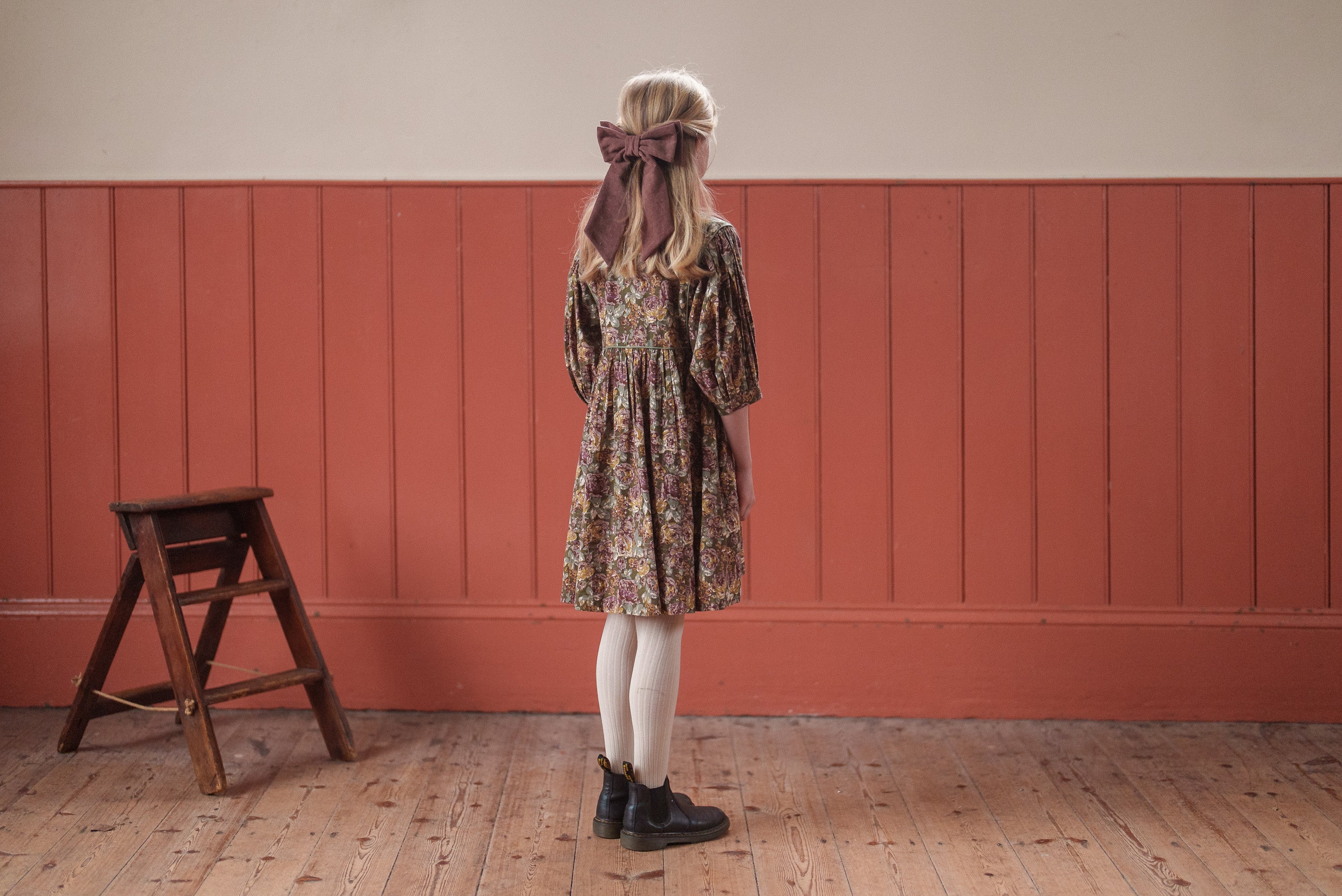 Young girl wearing an organic cotton vintage-style floral dress in green, styled with white tights, black shoes, and a large brown hair bow, standing on a wooden floor.