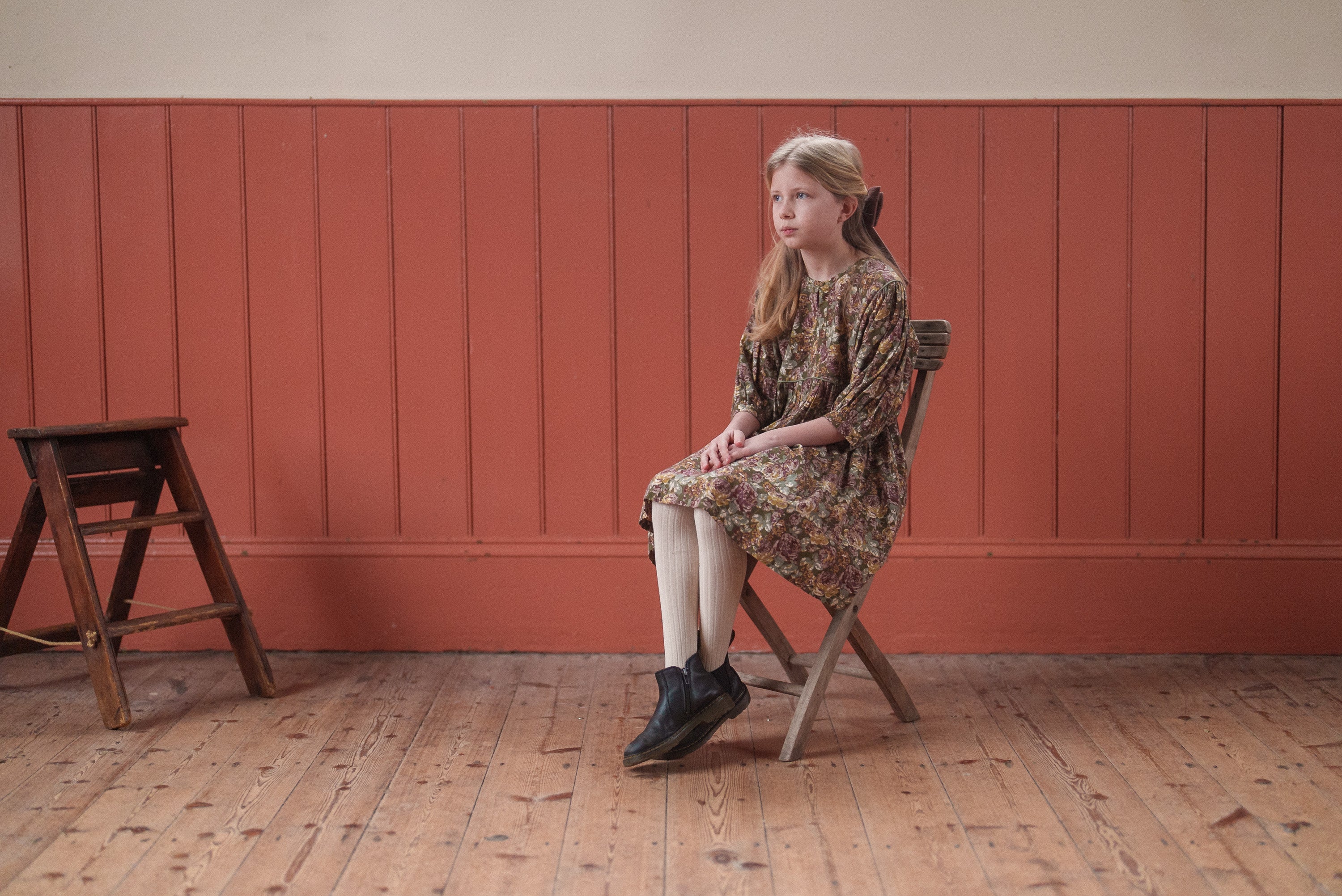 Young girl wearing a vintage style green floral dress made from organic cotton, sitting on a wooden chair against a warm-toned paneled wall in a natural light setting.