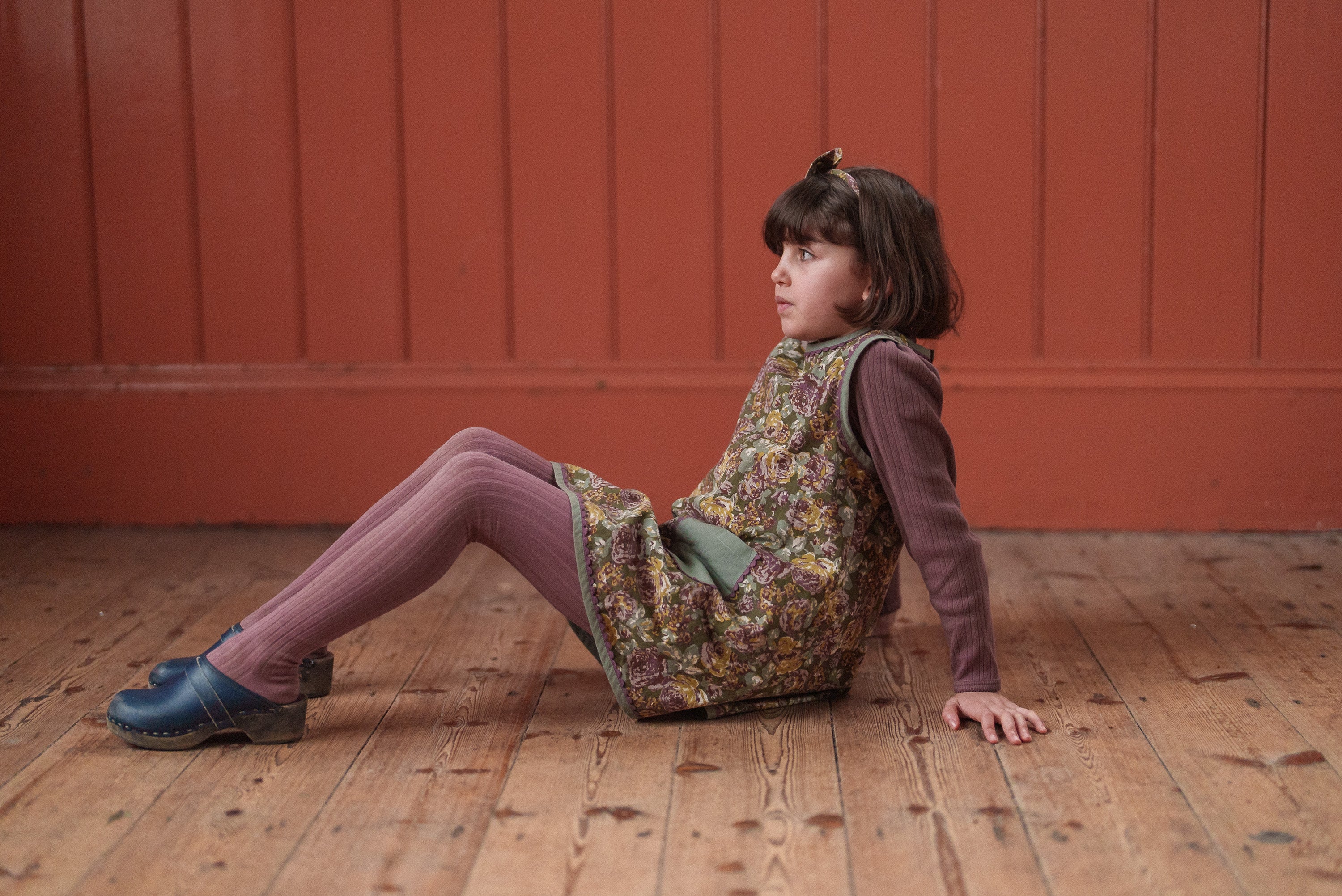 Young girl wearing vintage style floral dress in organic cotton sitting on wooden floor against a warm red wall, showcasing sustainable children's clothing.