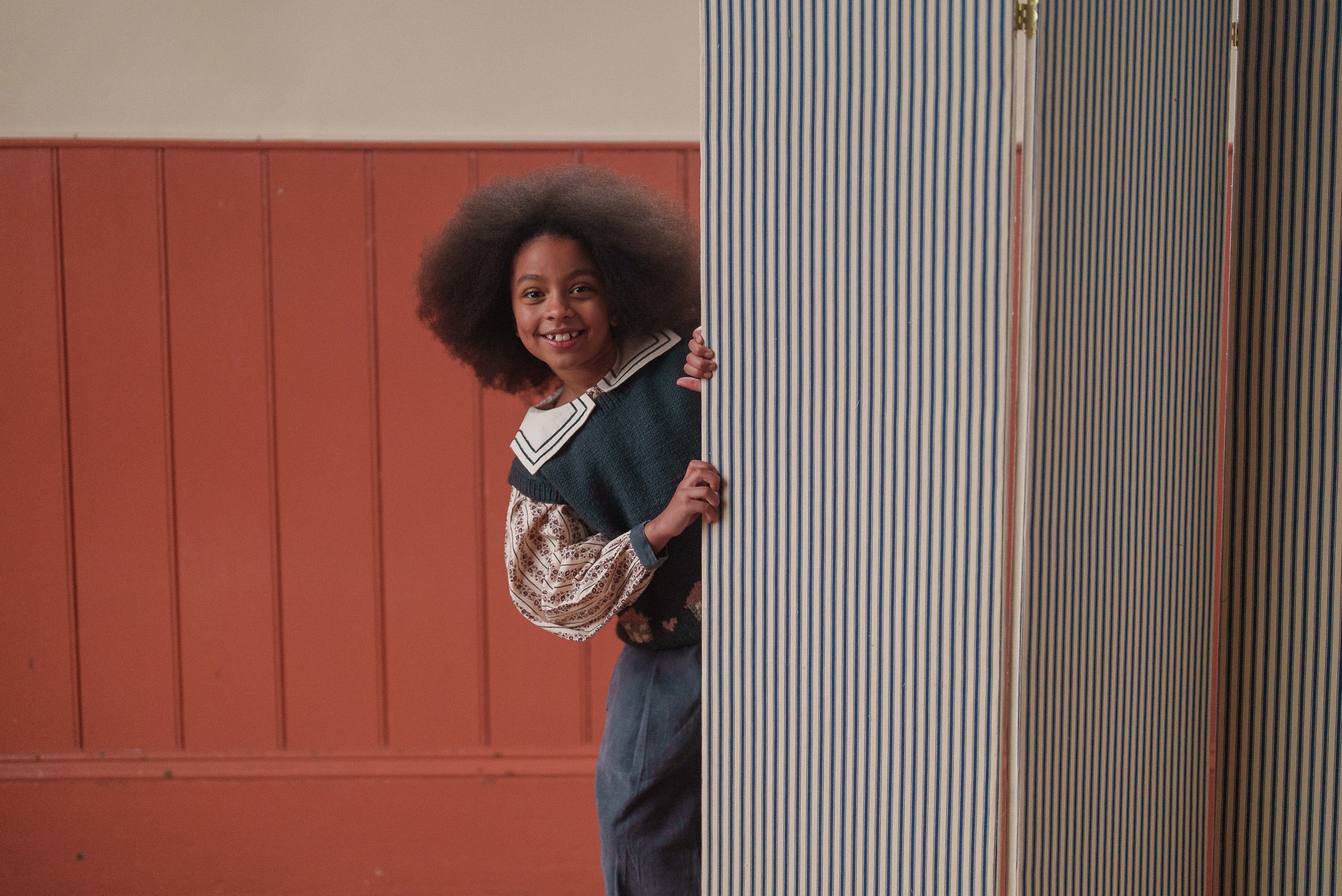 Smiling child wearing a multicolor merino hand-knitted vest peeks from behind striped curtains in a warm, vintage-inspired room, showcasing sustainable kids clothing.