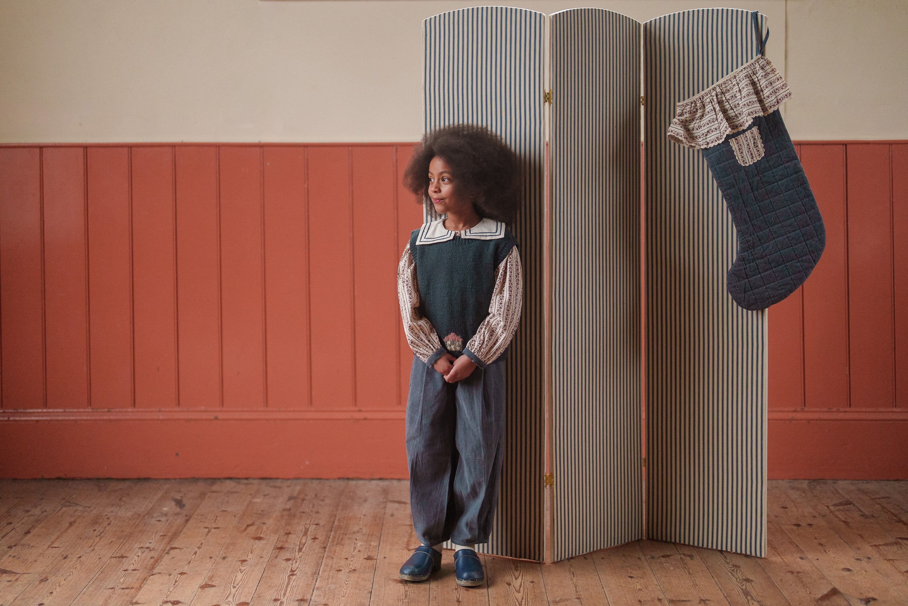 Young child wearing vintage style kids clothing with a hand-knitted multicolor merino vest, standing in a cozy room with natural wooden floors and warm tones.