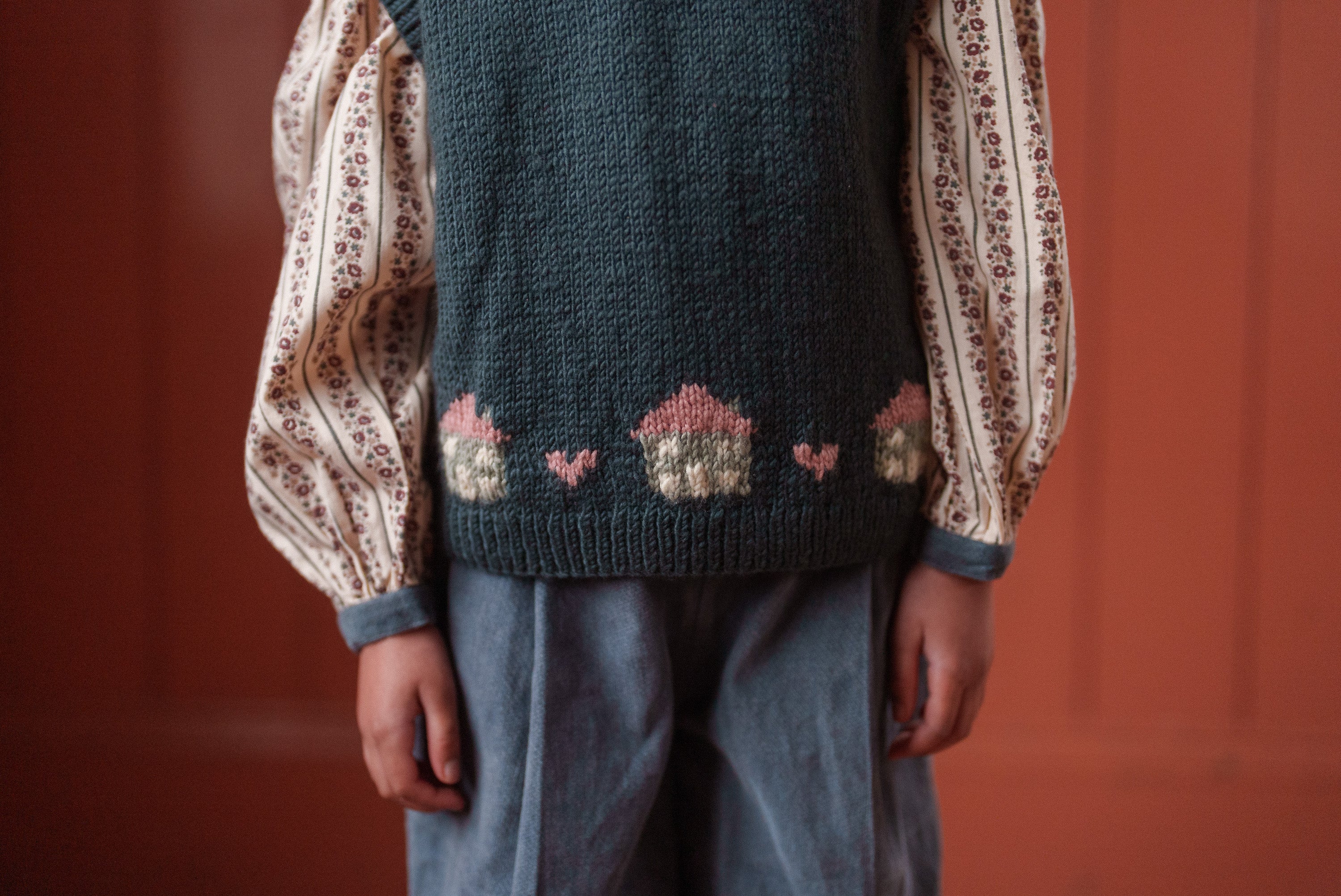 Child wearing a multicolor hand-knitted merino vest with cottage motifs over a vintage-style blouse and loose trousers, showcasing sustainable, ethically made children’s clothing.