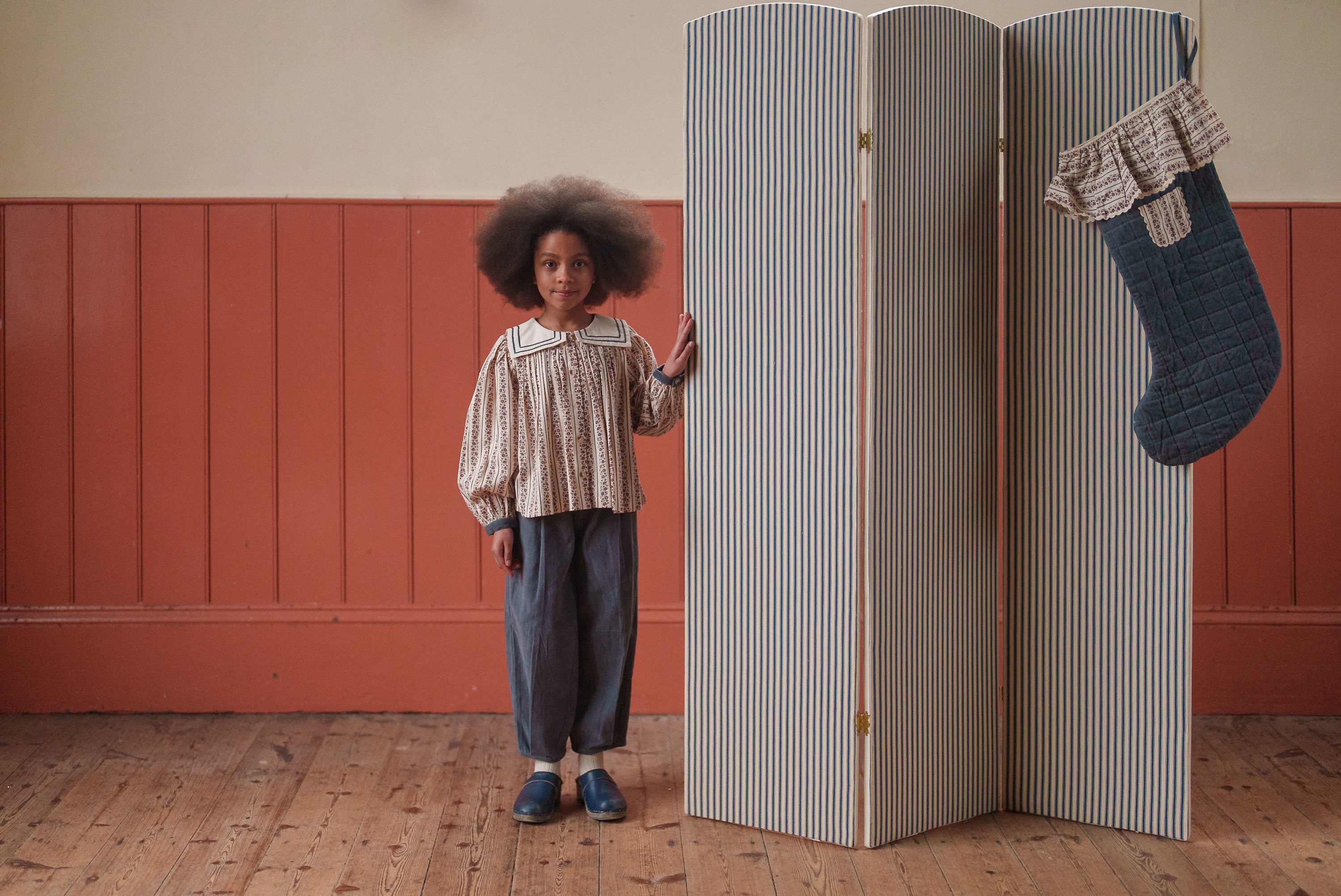 Child wearing vintage style girls blouse and blue velvet trousers made from organic cotton, standing beside a striped folding screen and hanging stocking in a warm, natural room.