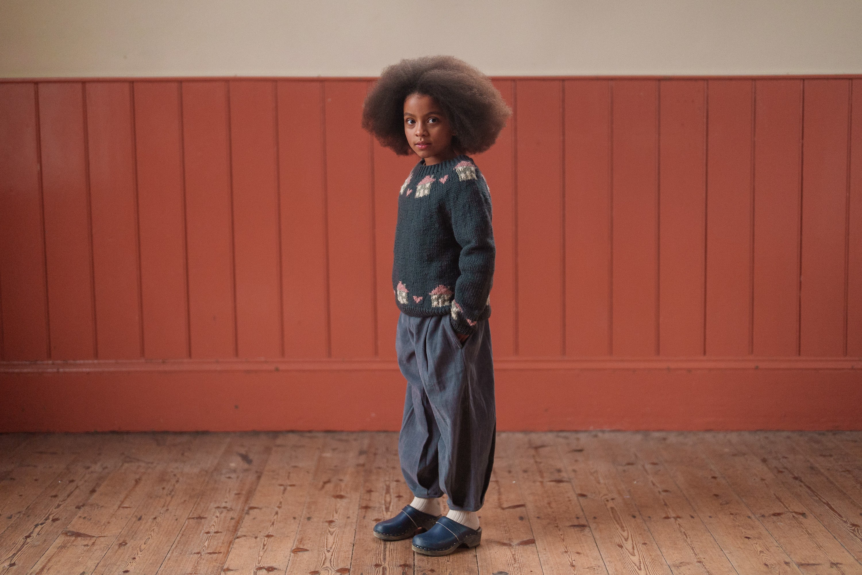 Child wearing a multicolor hand-knitted wool jumper and loose vintage-style pants, standing indoors on wooden flooring against a warm-toned wall.