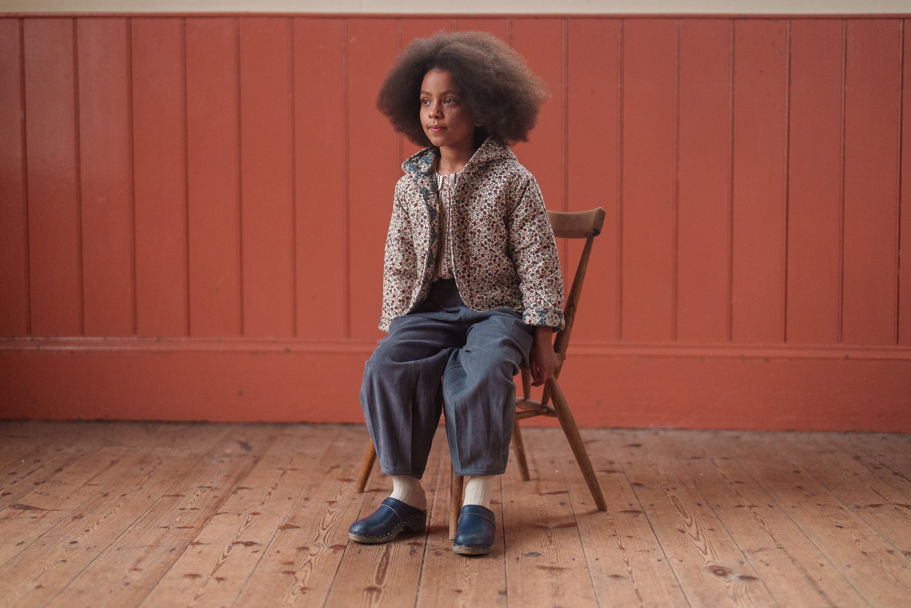 Child wearing vintage style children's clothing with blue velvet trousers made from organic cotton, sitting on a wooden chair against an orange wall.
