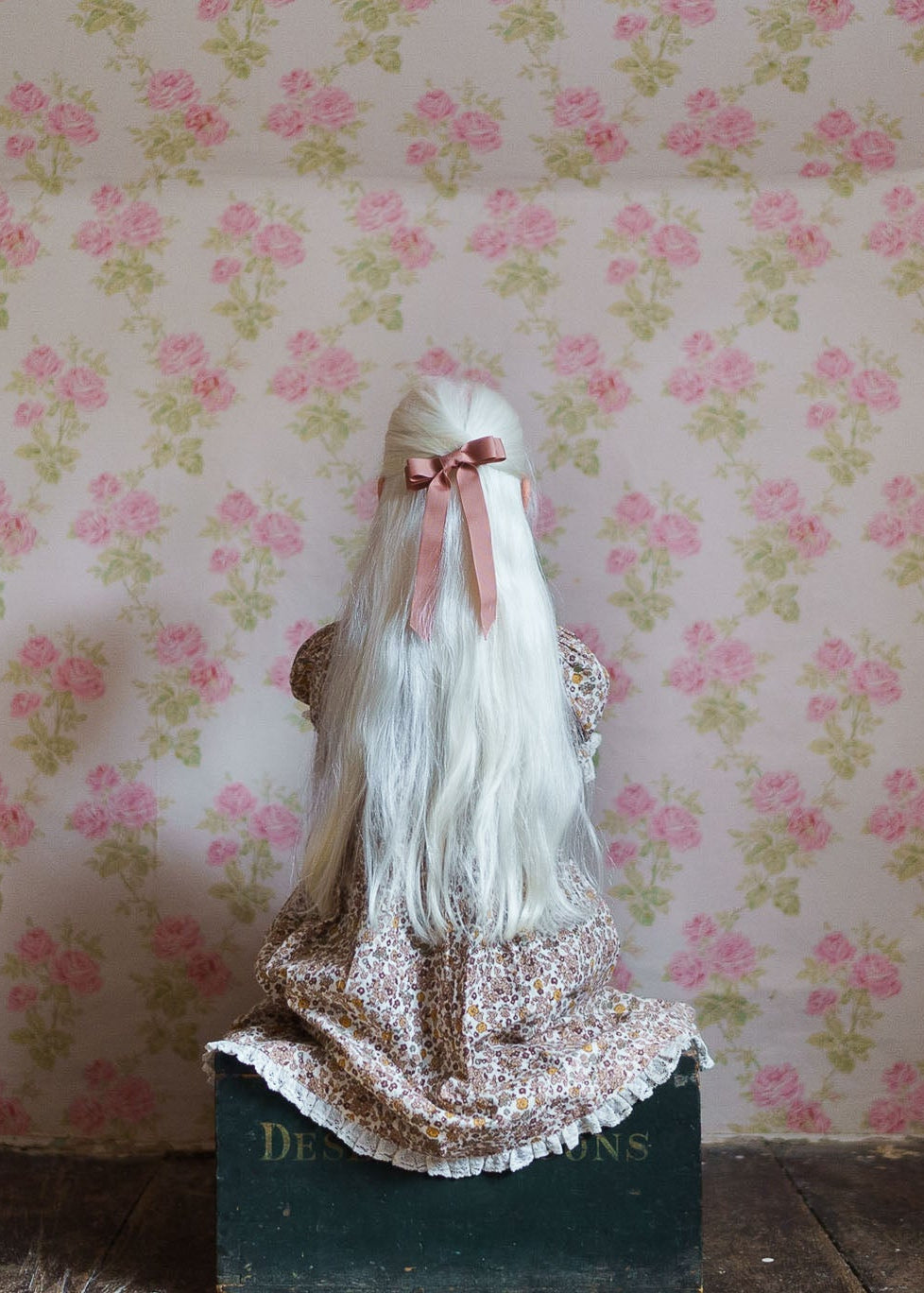 Child wearing a vintage style floral cotton blend dress with long white hair tied with a pink ribbon, sitting against a soft floral wallpaper background.