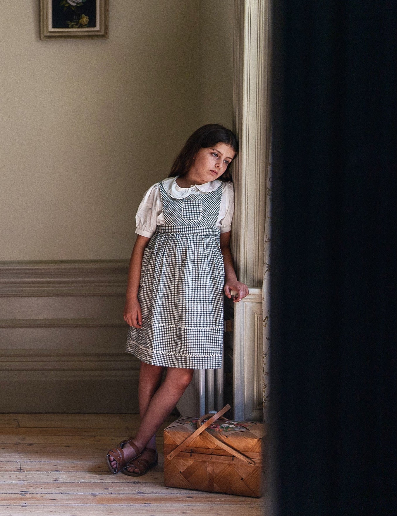 Young girl wearing a vintage style organic cotton blouse in off-white with puffed sleeves, paired with a checked dress, standing thoughtfully by a window in a softly lit room.