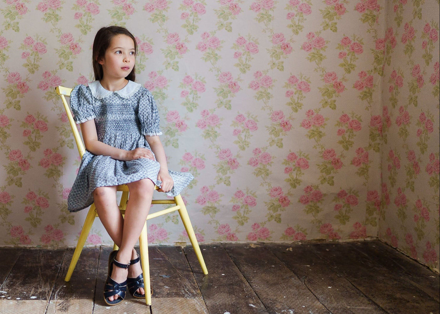 Young girl wearing a vintage-inspired blue floral smocked dress made from a cotton blend, sitting on a yellow chair against a floral wallpaper backdrop, showcasing sustainable children's clothing.
