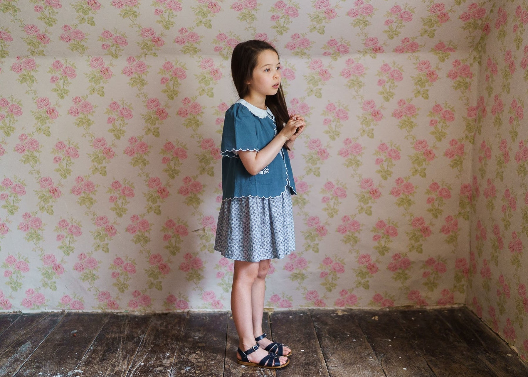 Young girl wearing a blue floral cotton blend vintage-style dress with smocked details, standing against a pink floral wallpaper backdrop in a natural light setting.