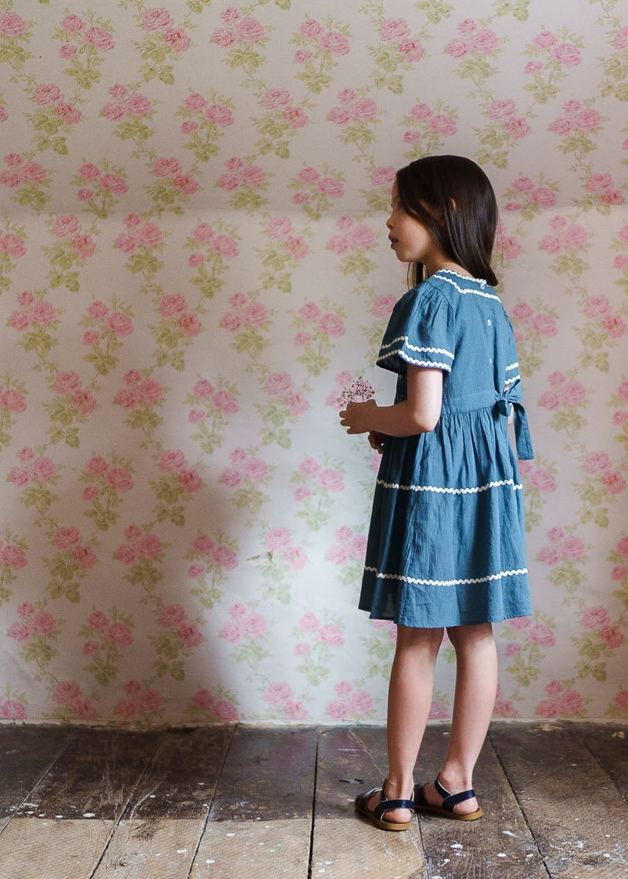Young girl wearing a vintage style blue smocked dress made from organic cotton, standing on wooden floor against a floral wallpaper, embodying natural and ethical children’s clothing.