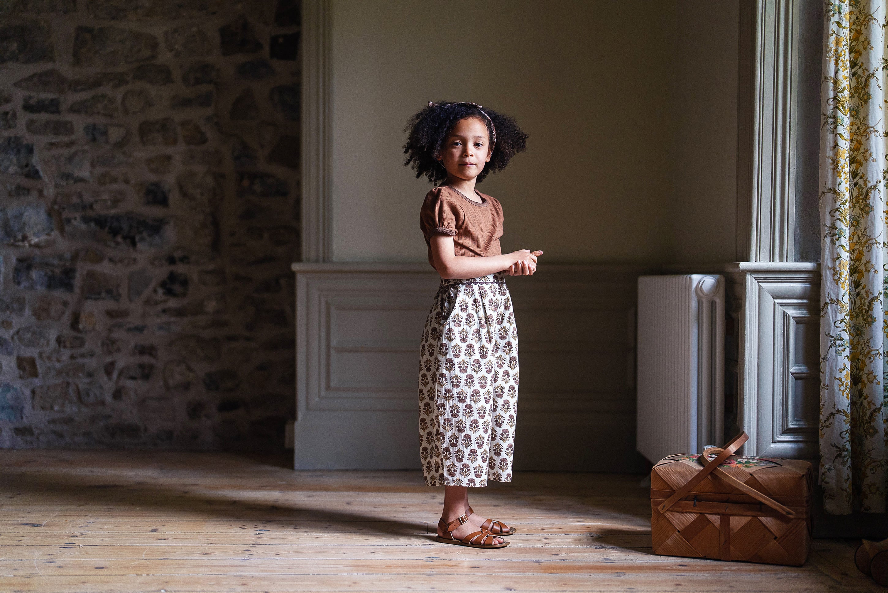 Young girl wearing a cinnamon-colored organic cotton pointelle T-shirt and vintage-style patterned skirt, standing in a softly lit room with natural materials and wooden floor.