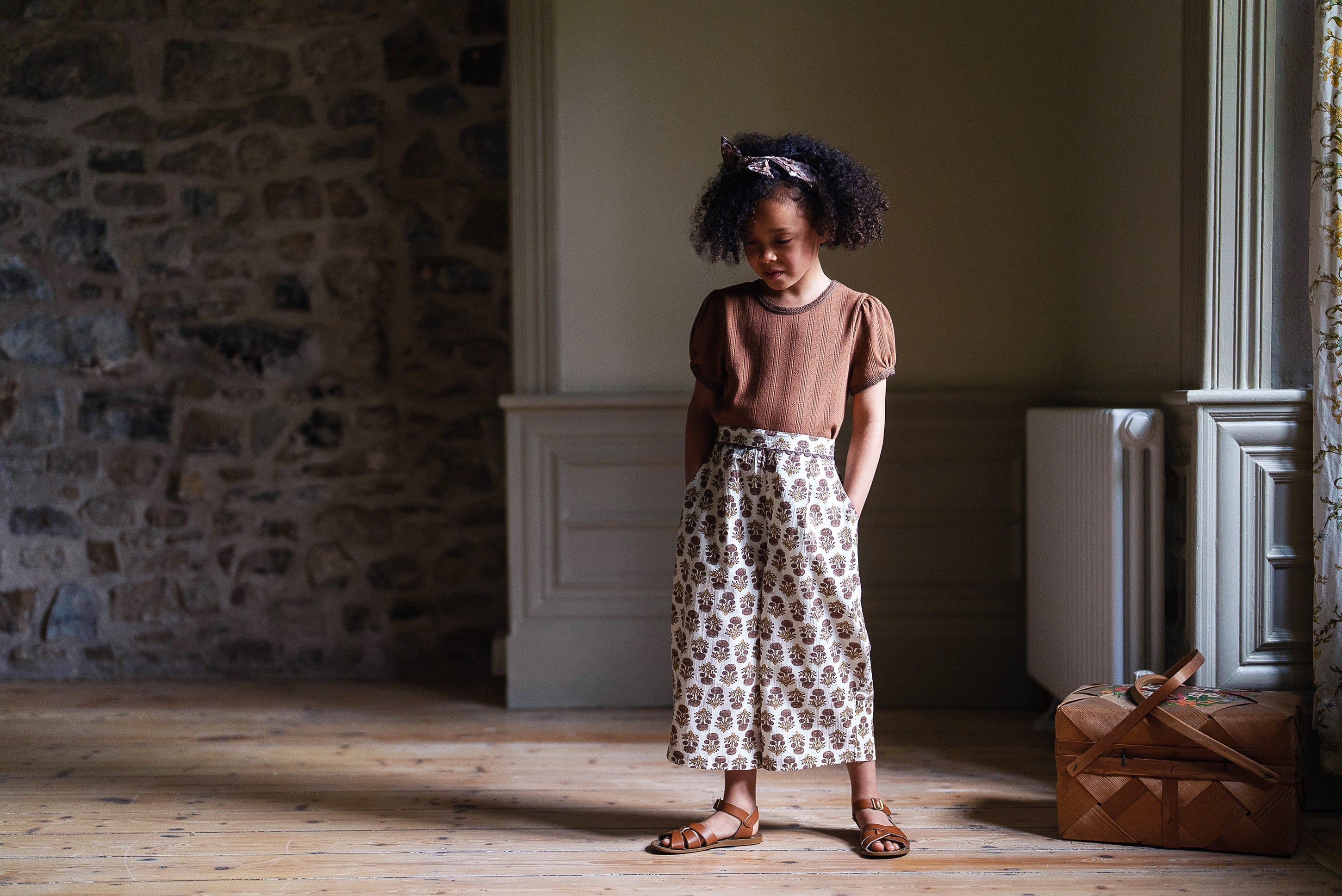 Young child wearing a cinnamon-colored organic cotton t-shirt with patterned vintage-style trousers, standing indoors on wooden floor in natural light, showcasing sustainable kids clothing.