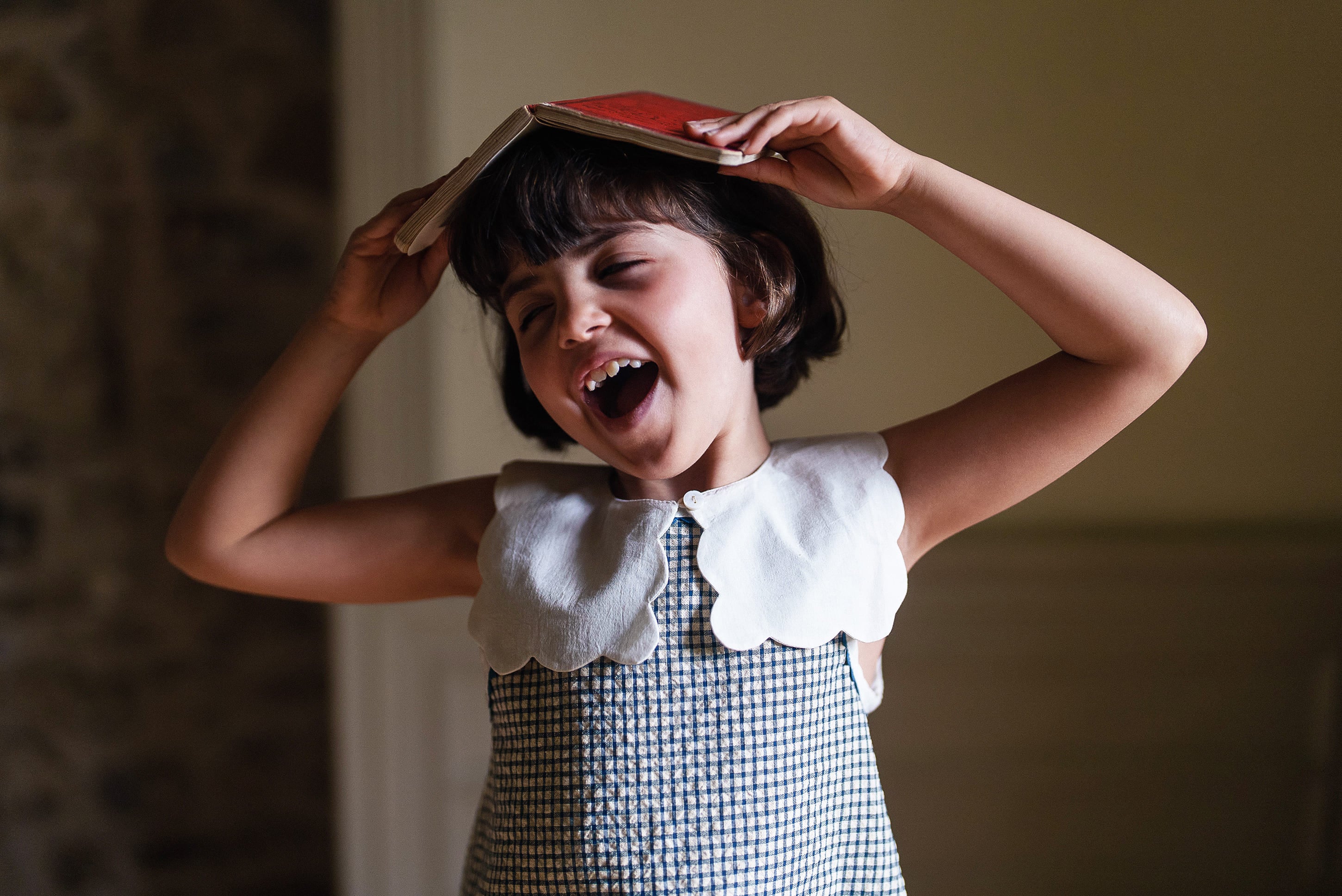 Happy child wearing vintage style dress with scalloped off white cotton collar, holding a book above their head in a warm, natural light setting.