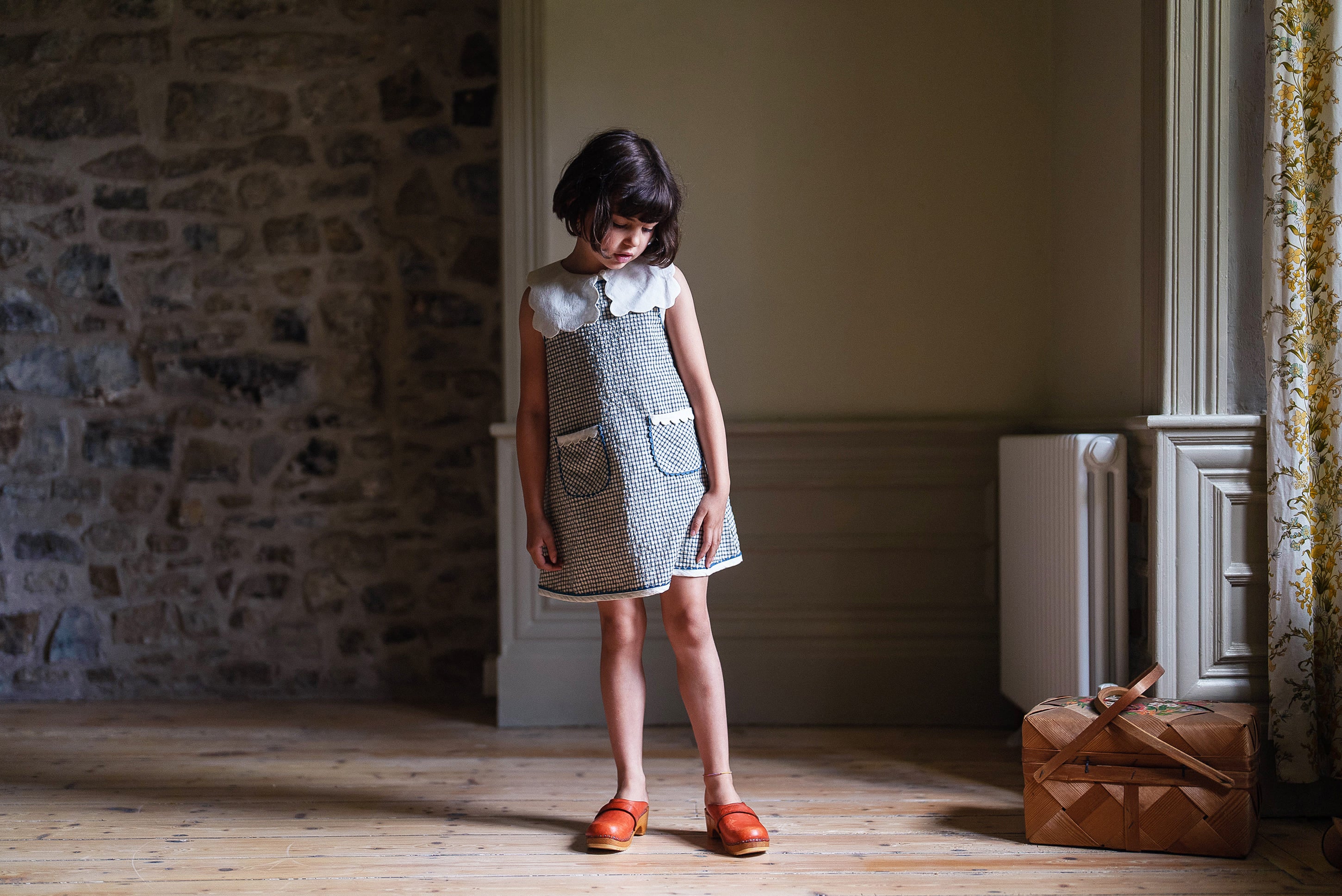 Young girl wearing a vintage style off white cotton dress with scallop collar, standing in softly lit room with wooden floor and stone wall, showcasing natural children's clothing.