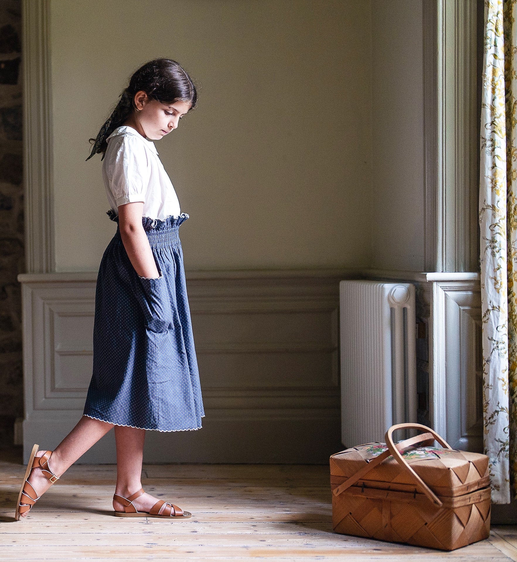 Young girl wearing an off-white organic cotton blouse paired with a vintage-style navy skirt, standing thoughtfully by a window in a softly lit room.