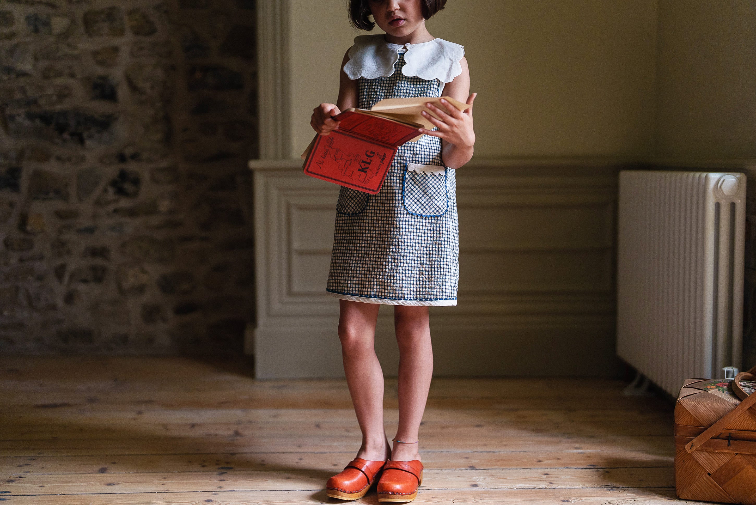 Young girl wearing a vintage style cotton dress with a scalloped white collar, standing indoors on wooden flooring, holding a book with a warm, natural light ambiance.