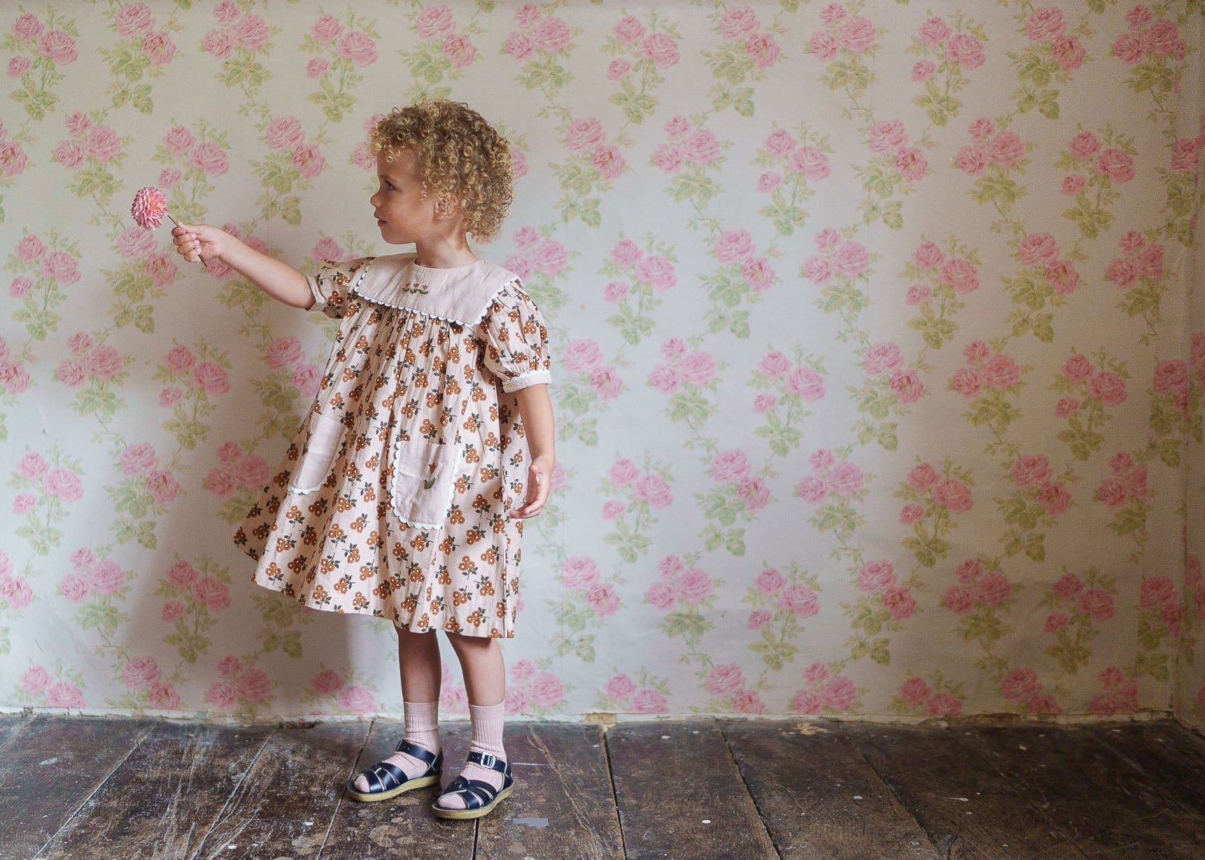 Toddler girl wearing a sustainable organic cotton floral dress with a vintage-inspired design, standing on wooden floor against a floral wallpaper background.