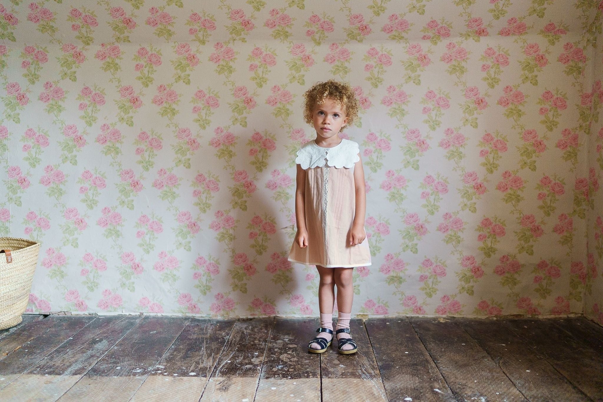 Young child standing in a powder pink organic cotton vintage style dress with a white collar and sandals, against a floral patterned wall in a rustic room.