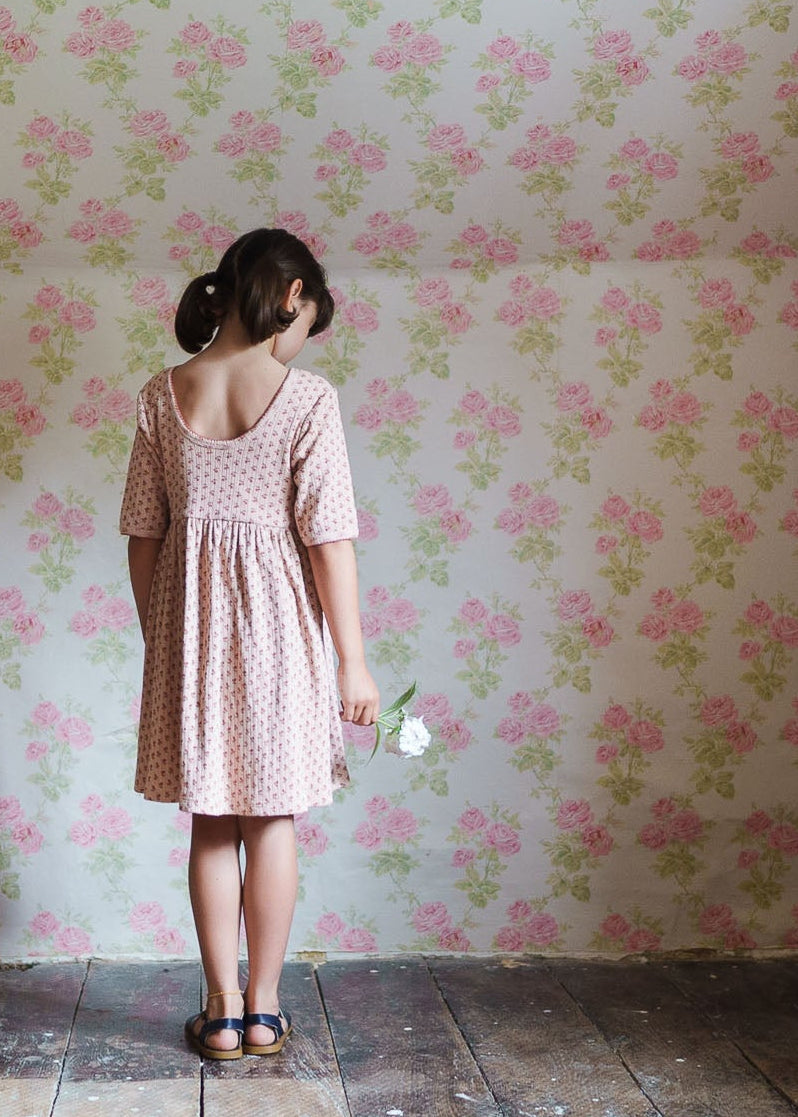 A young girl in a pink floral organic jersey cotton dress stands holding a white flower against a vintage-style floral wallpaper, showcasing natural, sustainable children’s clothing.