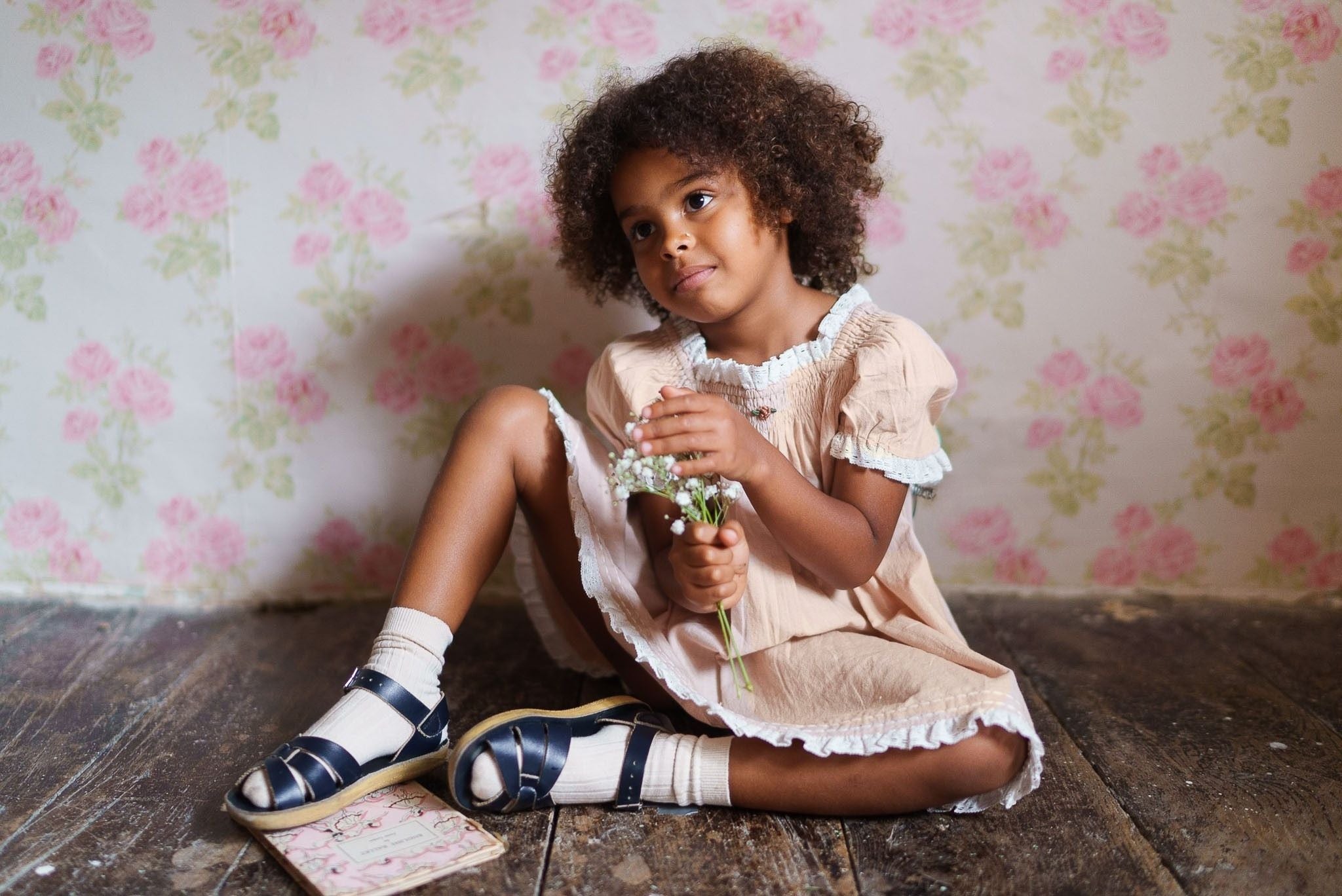Young girl wearing a powder pink organic cotton dress with vintage-inspired detailing, sitting on a wooden floor holding small white flowers in a softly lit room.