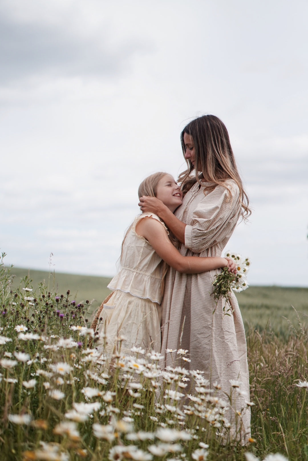 Two women embracing in a field of wildflowers under a cloudy sky.