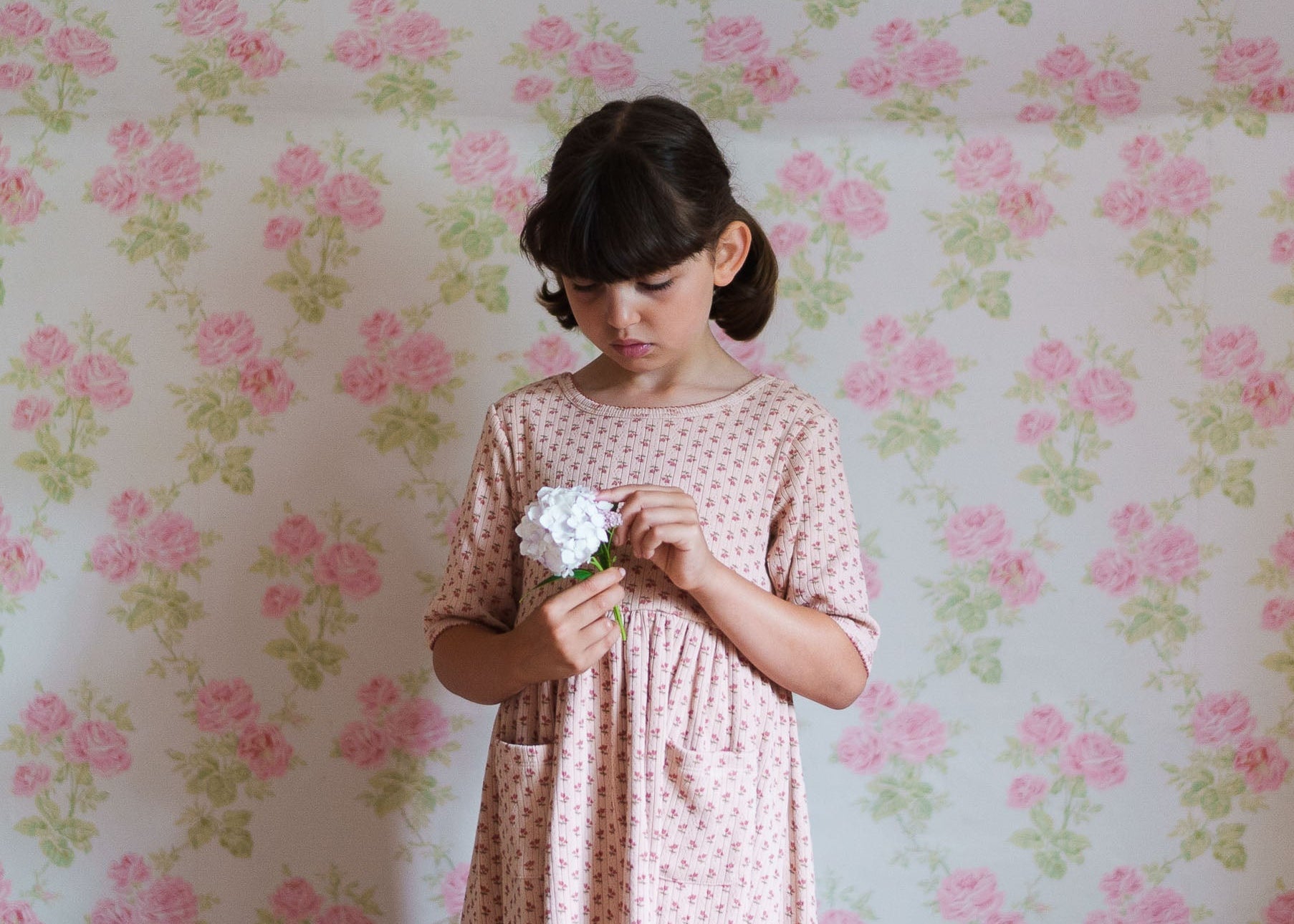 Young girl in a pink floral organic cotton dress holding a white flower, standing against a vintage-style floral wallpaper background.