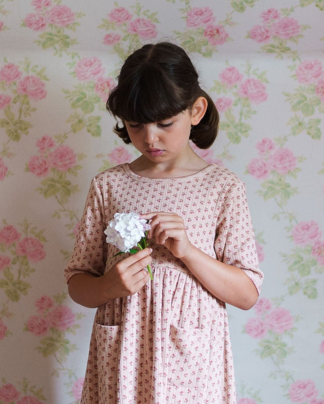 Young girl in a pink floral organic cotton dress holding a white flower, standing against a vintage-style floral wallpaper background.
