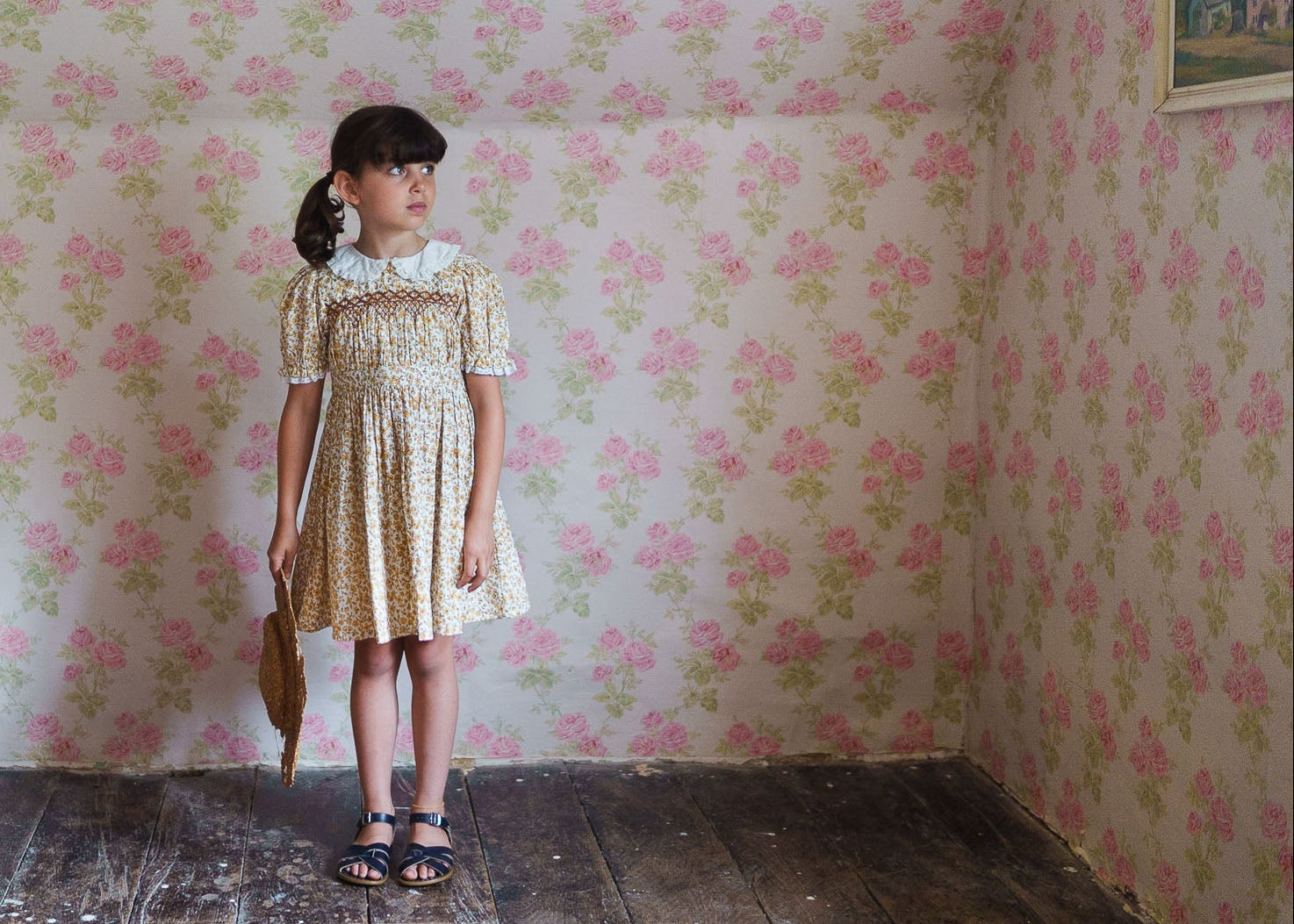 Young girl wearing a vintage-style floral cotton blend dress with a white collar, standing in a room with matching floral wallpaper, showcasing sustainable children's clothing.