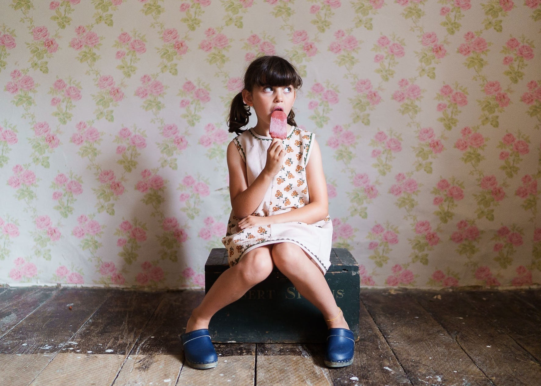 Young girl wearing a vintage-style floral dress made from organic cotton, sitting on a wooden floor with a nostalgic floral wallpaper background, enjoying a popsicle.