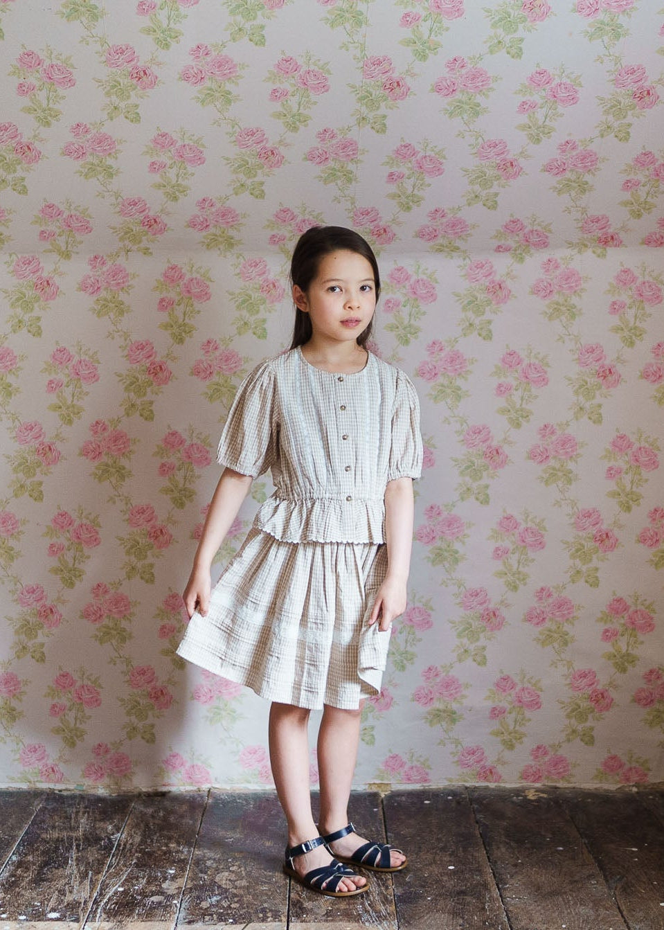 Young girl wearing a cream checkered organic cotton skirt and top set, standing against vintage floral wallpaper in a natural, sustainable children’s clothing style.