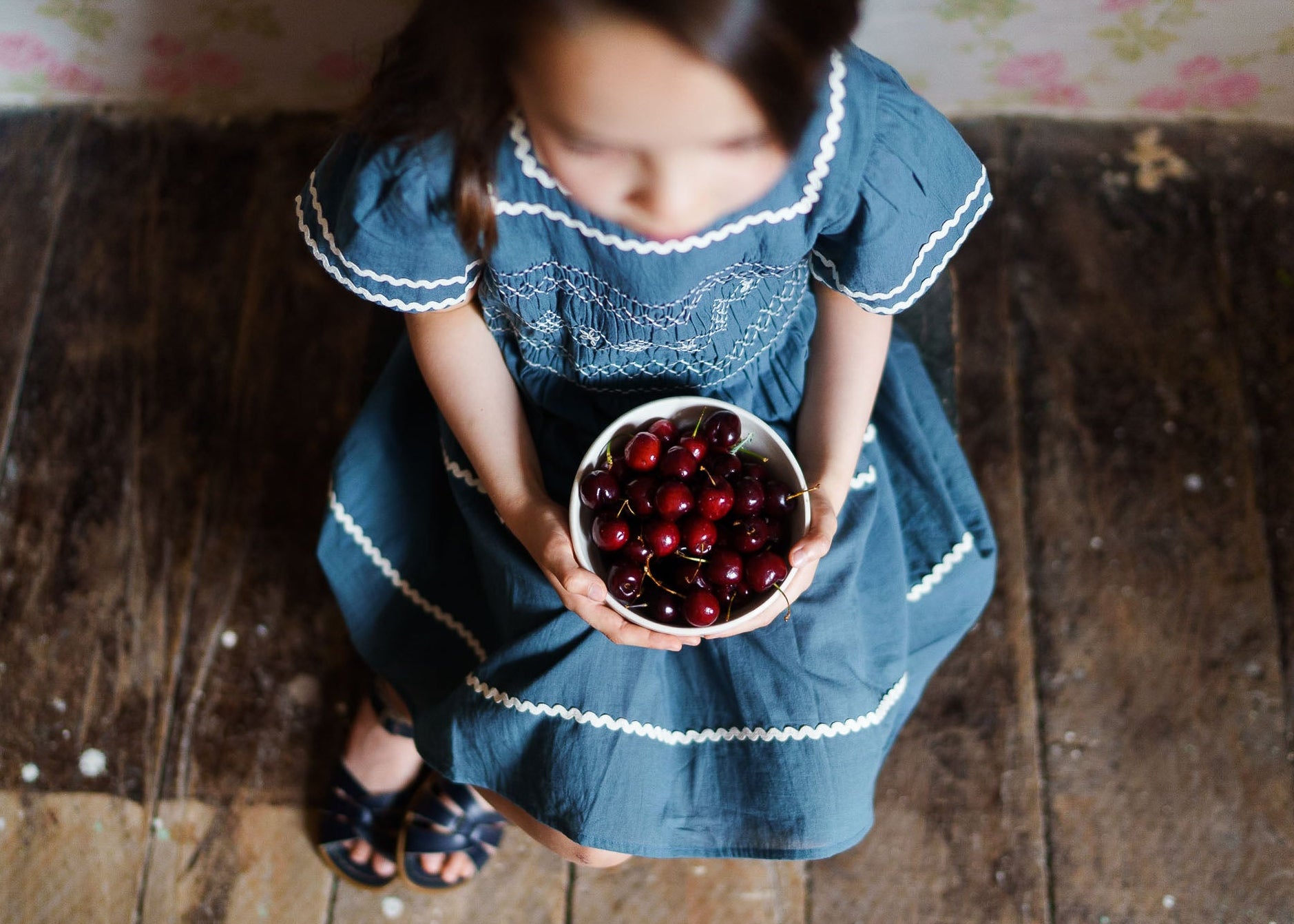 Young girl in a vintage-style blue organic cotton dress holding a bowl of cherries, showcasing sustainable, ethically made children's clothing with a natural feel.