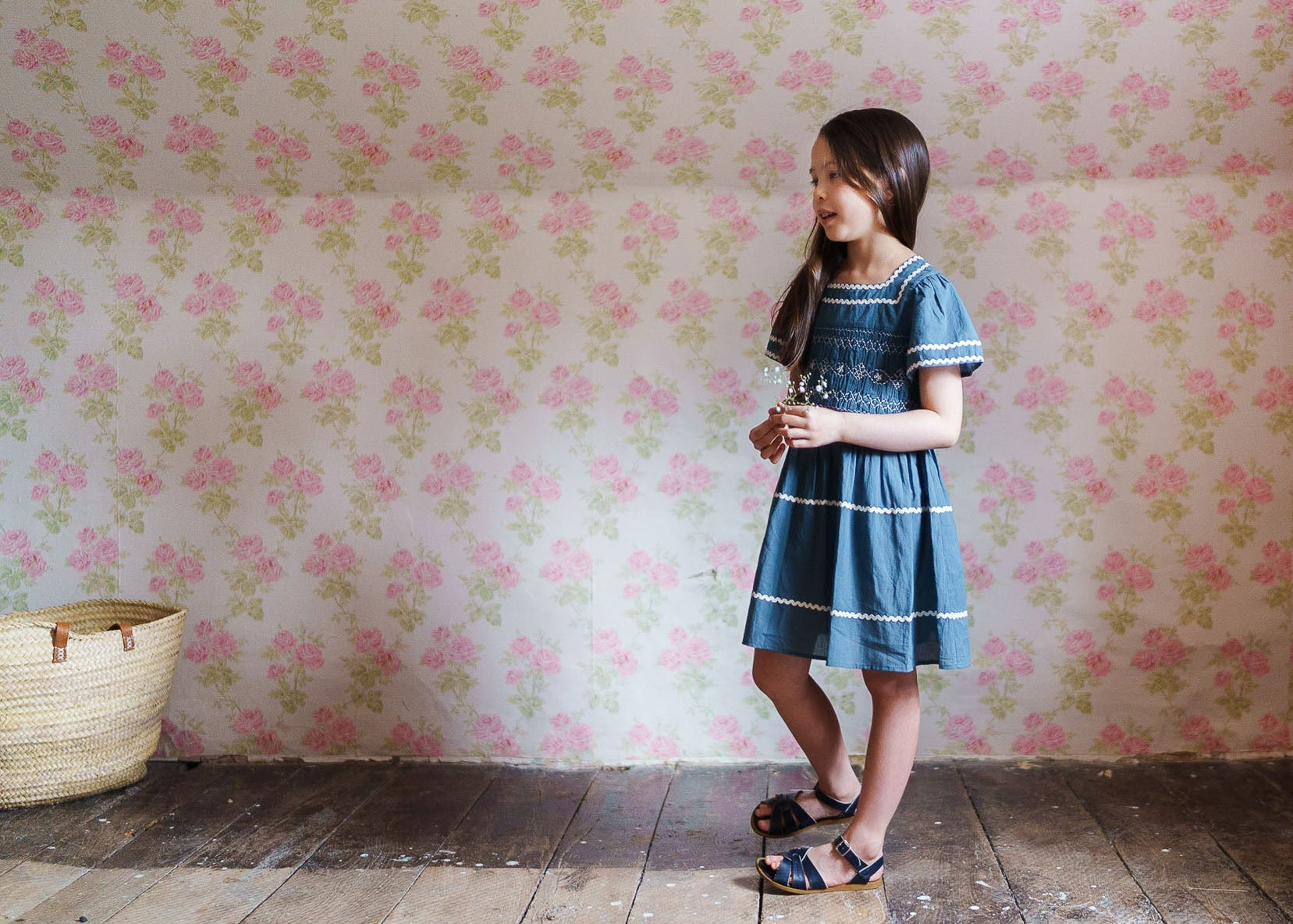 Young girl wearing a vintage style blue smocked dress made from organic cotton, standing on wooden floor against floral wallpaper in a natural, ethically made children's clothing setting.