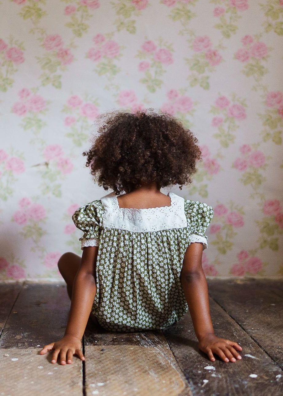 Child wearing a vintage style romper with a green floral pattern and white lace collar, sitting on a rustic wooden floor against a floral wallpaper, in organic children's clothing.