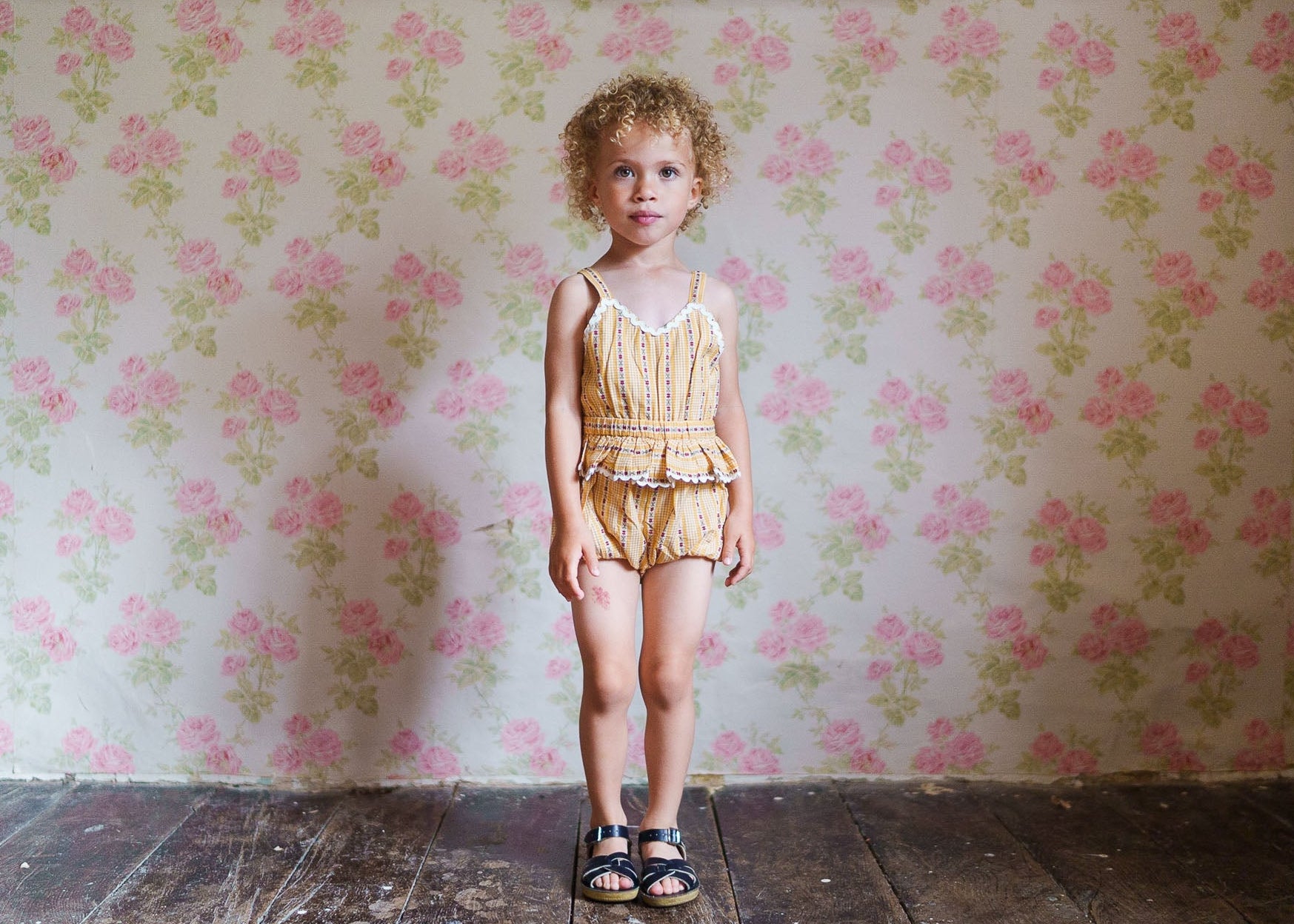 Young child wearing a vintage style romper with delicate floral and stripe patterns, standing against a floral wallpaper background, showcasing natural, organic children’s clothing.