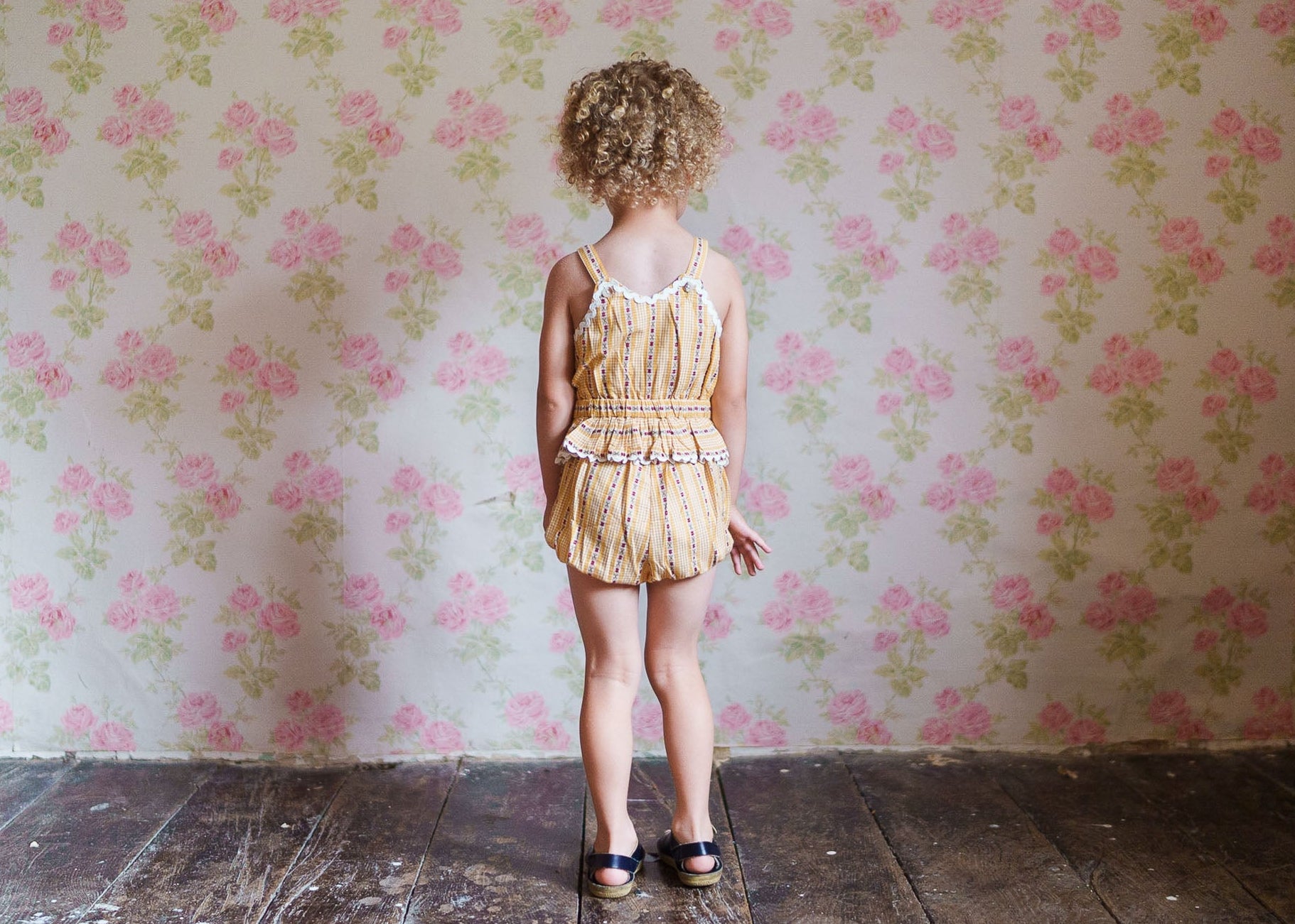 A young child wearing a vintage style romper with a delicate floral pattern, standing barefoot on a wooden floor against a pastel pink floral wallpaper background.