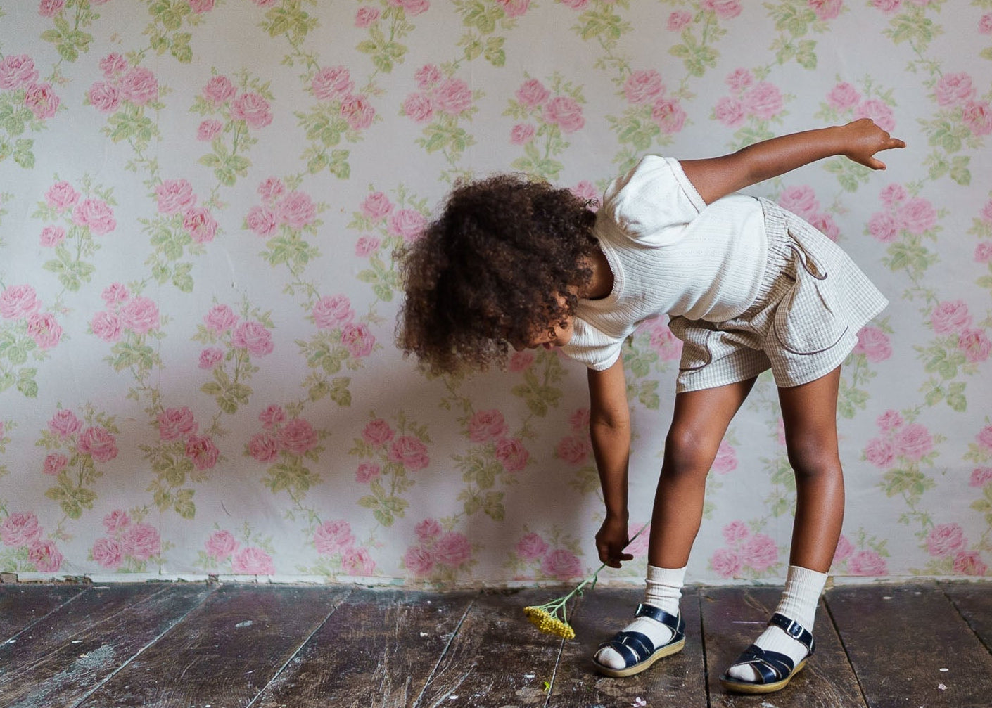 A young child wearing vintage-style natural cotton shorts with a straw check pattern, paired with a simple white top, playing indoors on a wooden floor.