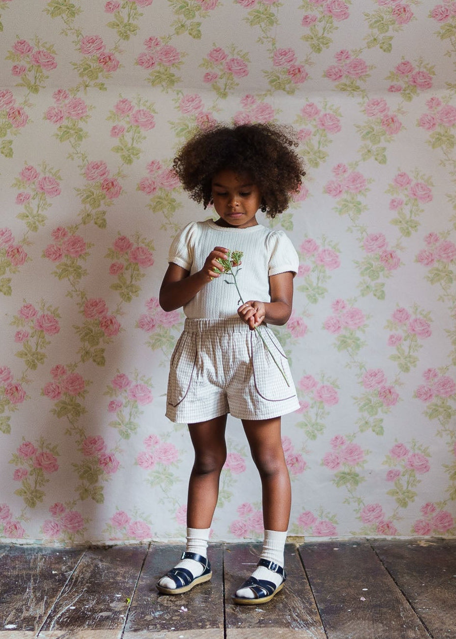 Young girl wearing vintage-style organic cotton shorts with a subtle check pattern, standing against floral wallpaper, holding small green flowers gently.