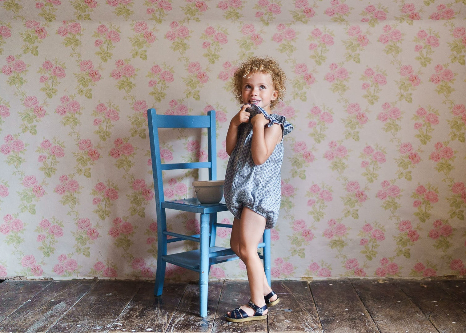 Young child wearing a vintage style romper with floral patterns, standing next to a blue wooden chair on rustic wooden flooring against a floral wallpaper background.