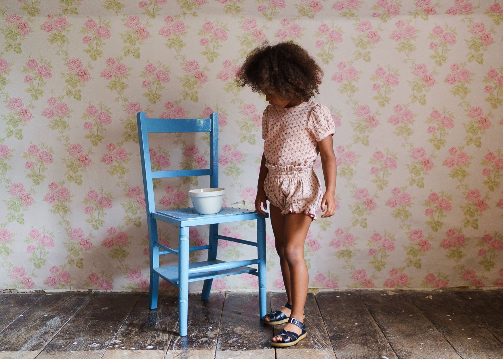 Toddler girl wearing pink floral organic cotton bloomers standing next to a blue wooden chair with a white bowl, in a vintage-style room with floral wallpaper.