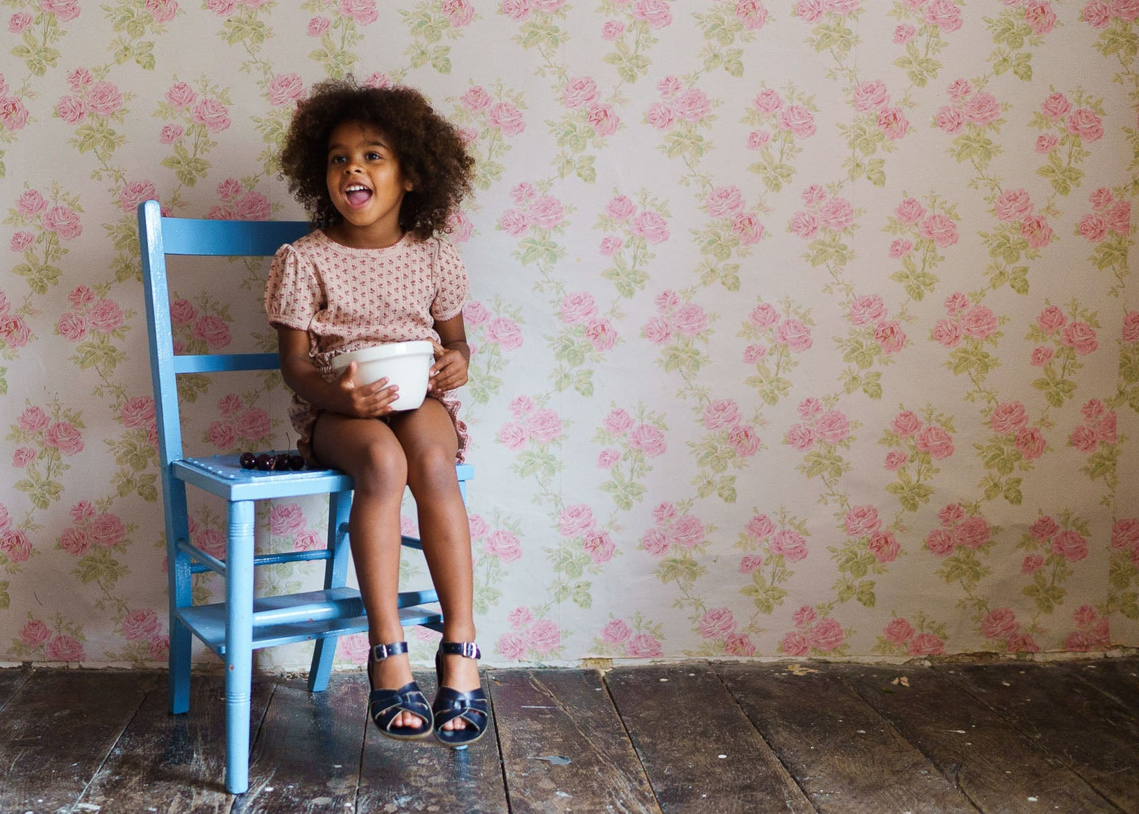 Smiling young girl wearing a pink organic cotton pointelle t-shirt with tulip floral pattern, sitting on a blue wooden chair against vintage floral wallpaper, showcasing sustainable children's clothing.