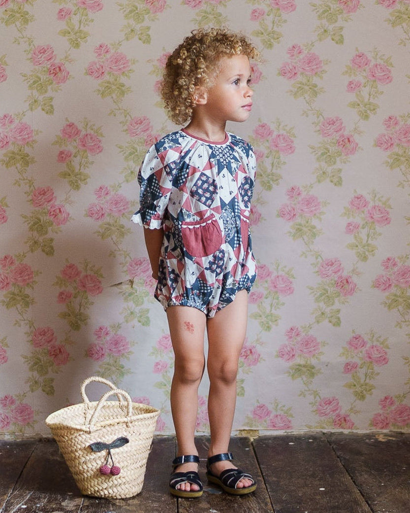 Young child wearing a vintage-style patchwork floral romper made from soft organic cotton, standing on wooden floor with a woven basket nearby, in a cozy, natural setting.