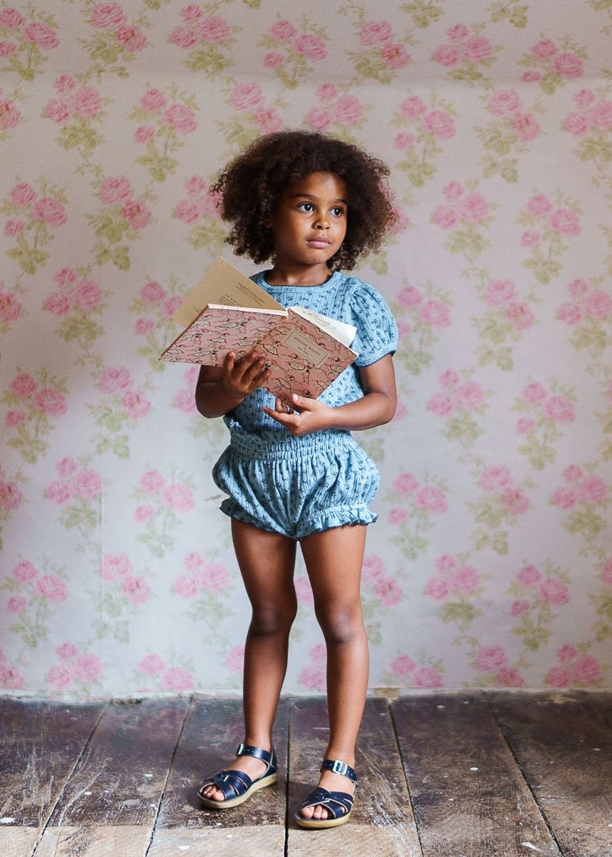 Young girl wearing blue floral organic cotton bloomers and top, holding vintage-style books, standing on wooden floor against floral wallpaper in a natural, vintage-inspired children’s clothing setting.
