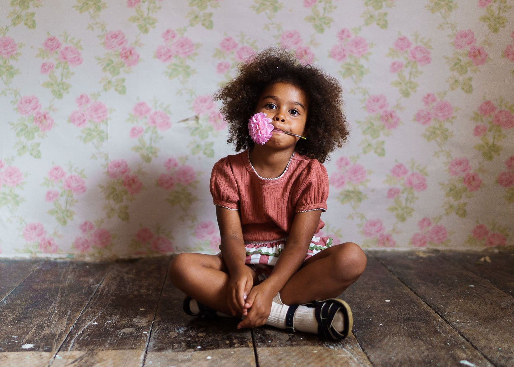Young child sitting on a wooden floor wearing a pink organic cotton top, holding a pink flower in their mouth, with a vintage floral wallpaper background.