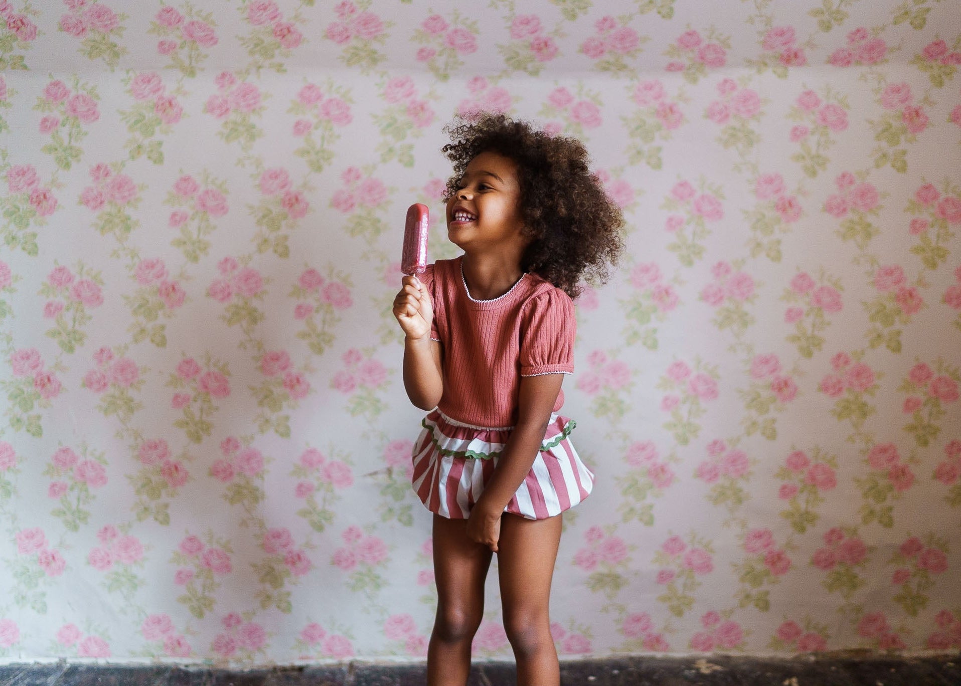 Happy young girl wearing a pink organic cotton top with vintage style striped shorts, enjoying a raspberry popsicle against a floral wallpaper background in a natural, ethical children’s clothing setting.