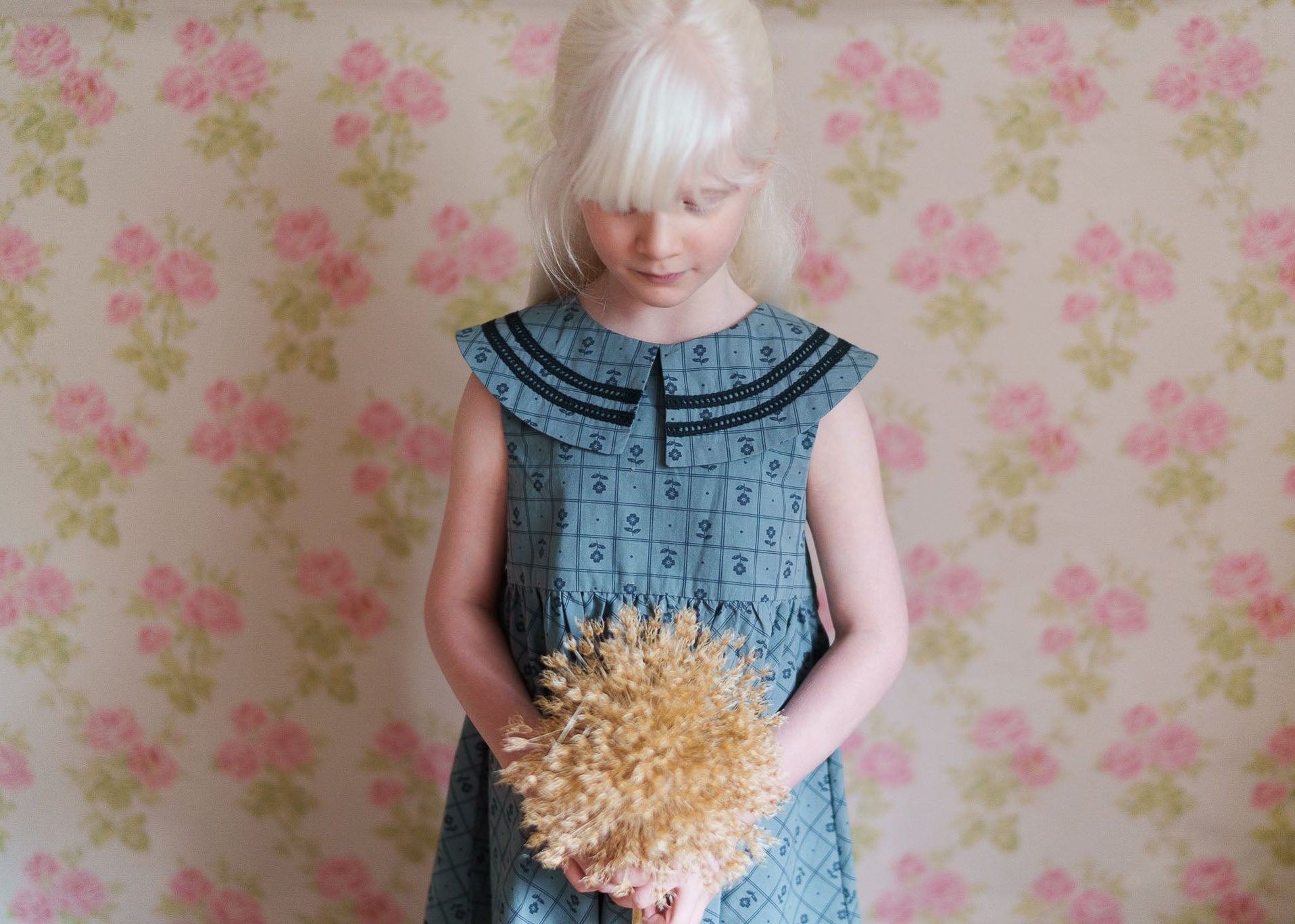 Young girl wearing a vintage style organic cotton dress with a floral pattern, holding dried flowers against a soft floral wallpaper background.